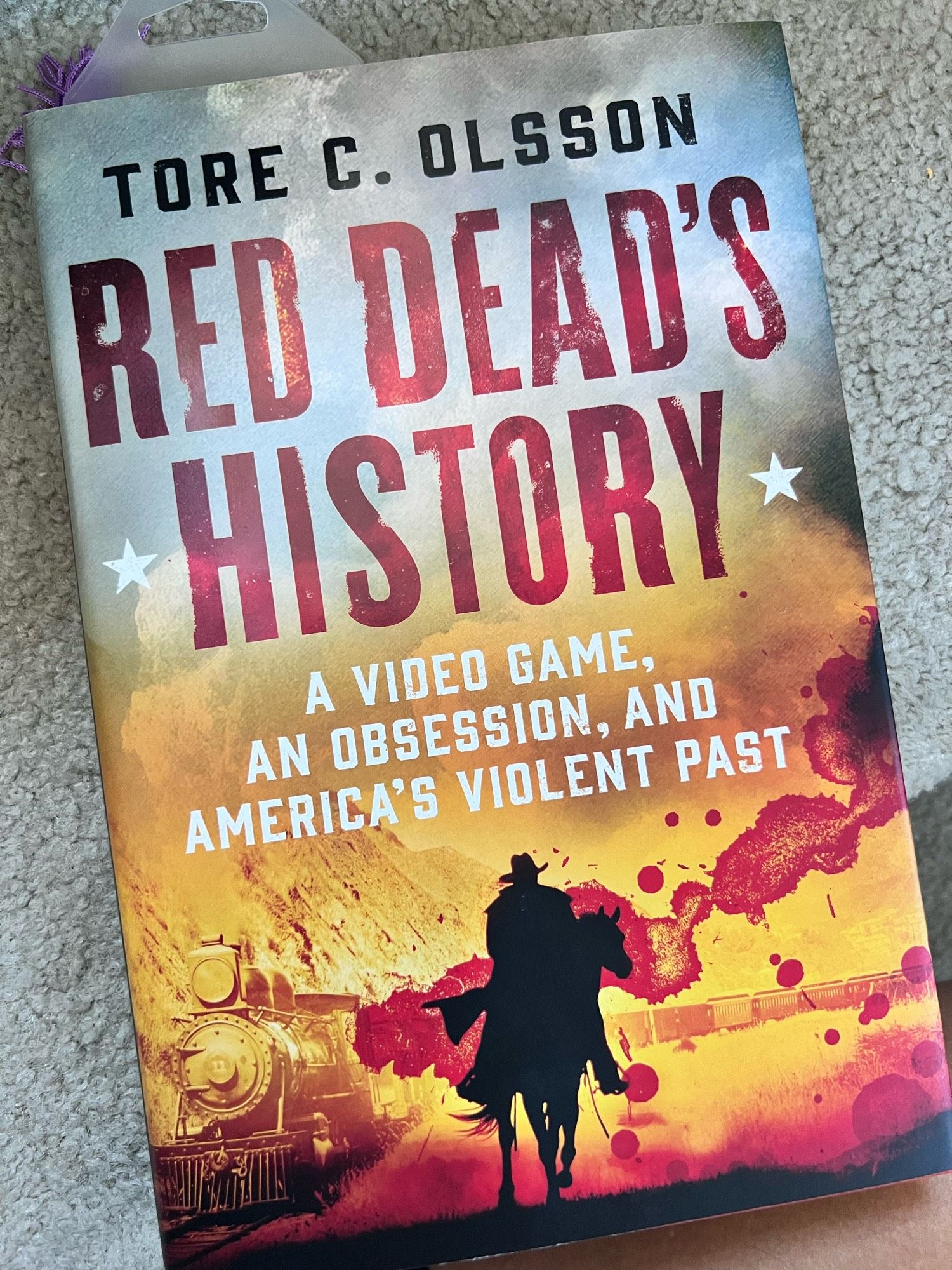 Photo of book “Red Dead’s History: a video game, an obsession, and America’s violent past” by Tore C Olssen, cover is a train coming through a mountain range with a blood splatter-esque red splotch and a black silouhette of Arthur Morgan on a horse from Rockstar Games Read Dead Redemption 2