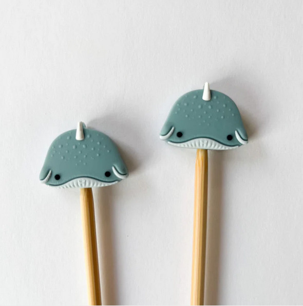 A pair of bamboo or wood single point knitting needles each have an adorable silicon narwhal needle point protector on top. 