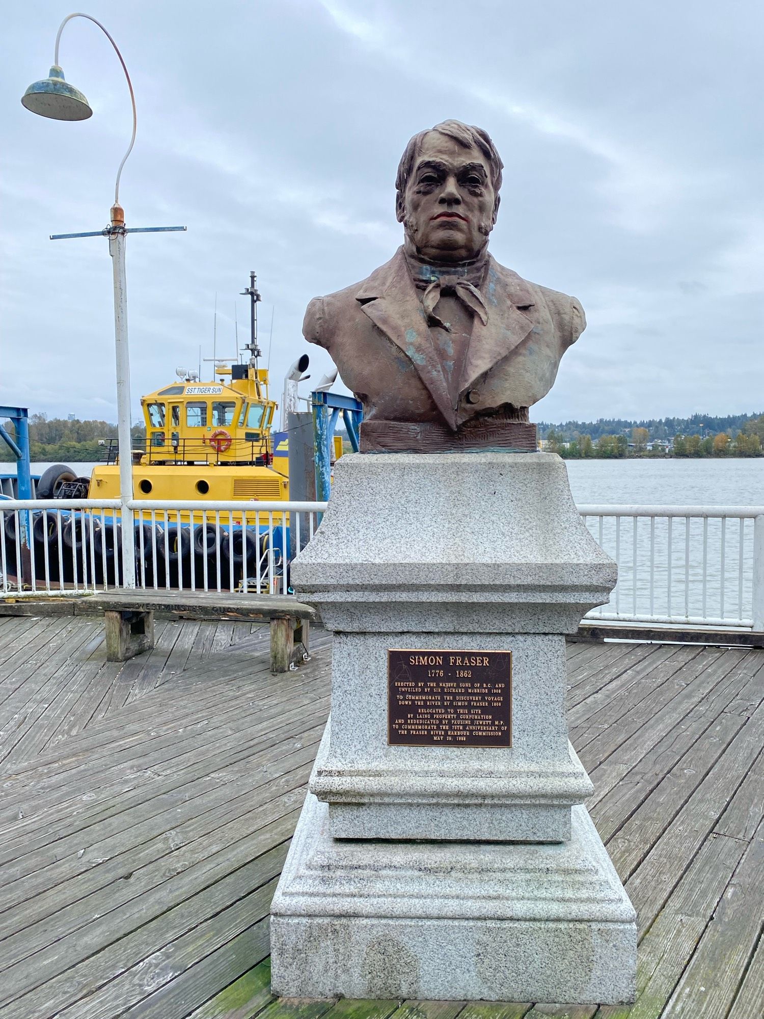 Bust of Simon Fraser on the boardwalk by the river with a yellow tugboat in the background