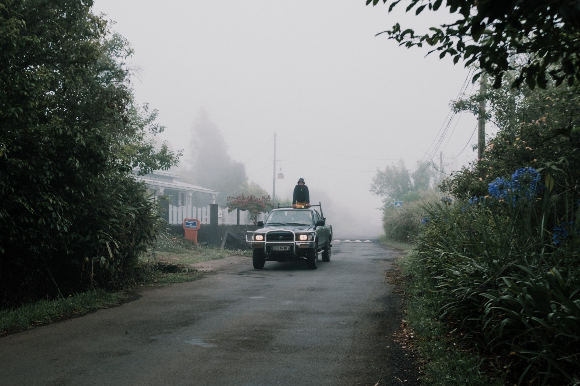 Un véhicule plus loin sur une route bordé de végétation sort de la brume. Un homme se tient debout sur l'arrière du pick-up