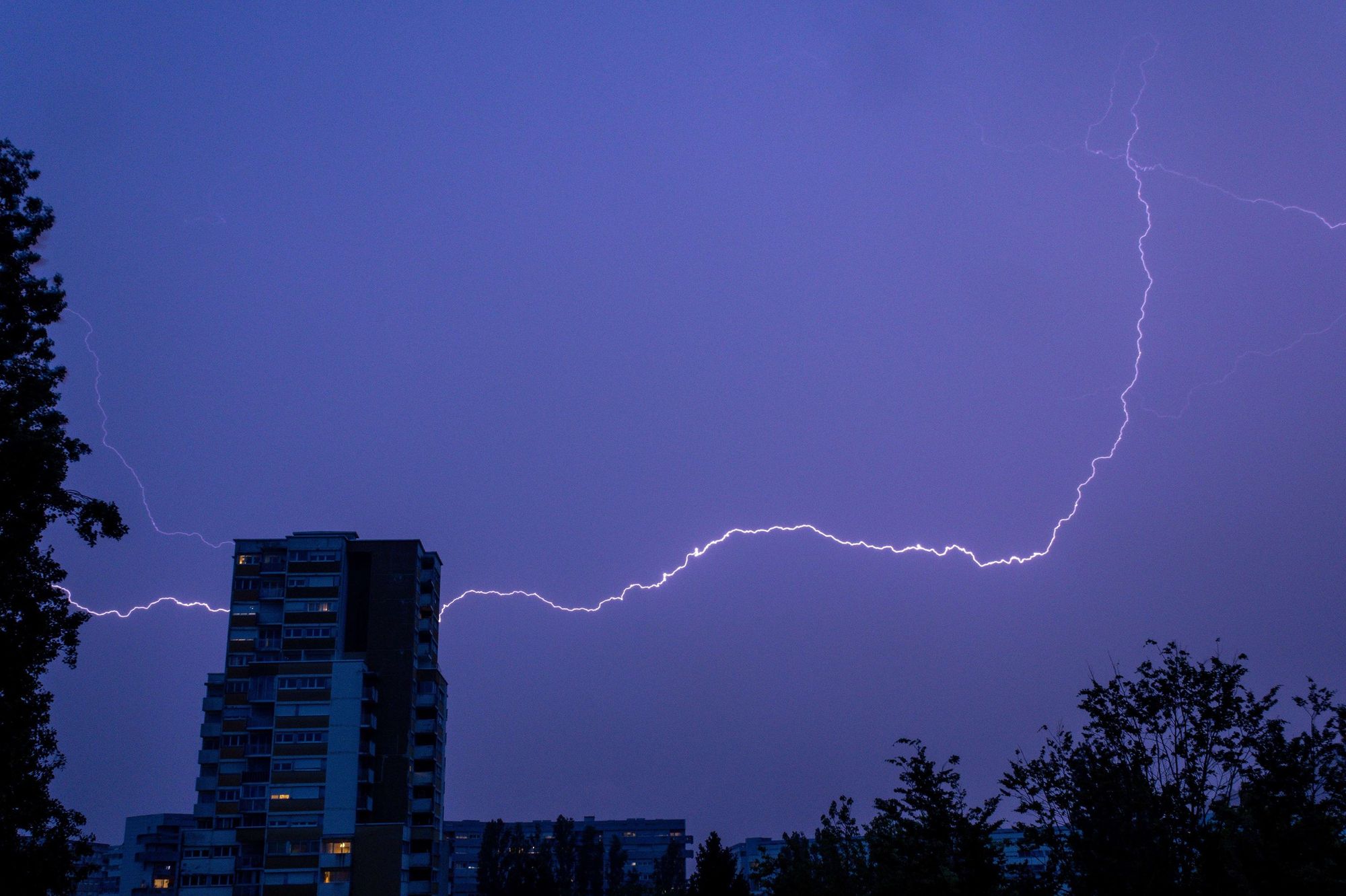 Photo d'un éclair nocturne qui passe derrière une tour d'habitation. Il semble entrer d'un côté et sortir de l'autre.