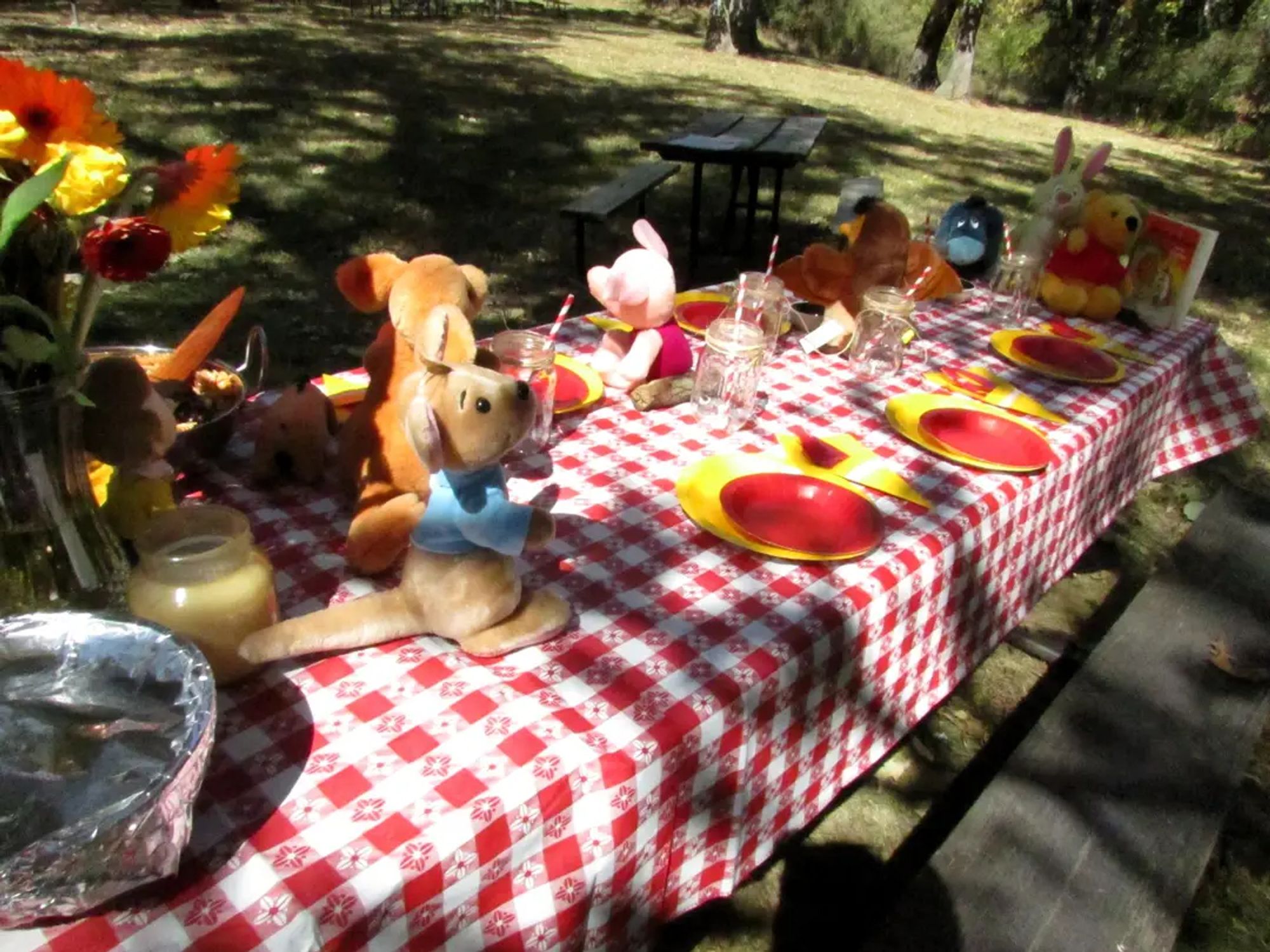 A picnic table with red/white checker tablecloth and stuffed animals of all the Pooh characters. There is a floral bouquet visible and red and yellow paper plates.