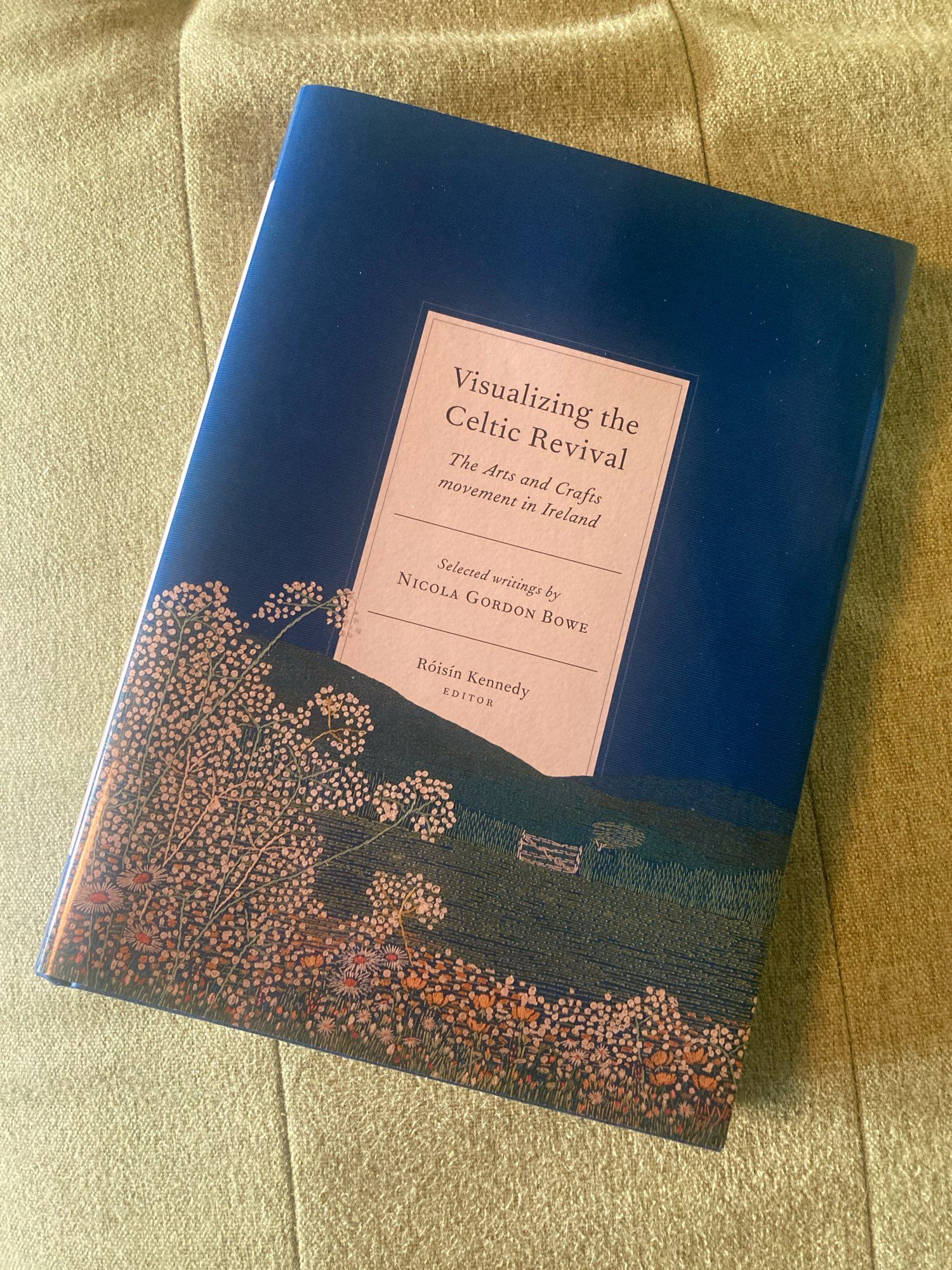 A hardback book with an embroidered image on the cover, blue sky, green grass and white flowers. Visualizing the Celtic Revival: The Arts and Crafts Movement in Ireland: Selected Writings by Nicola Gordon Bowe, Róisín Kennedy, editor.