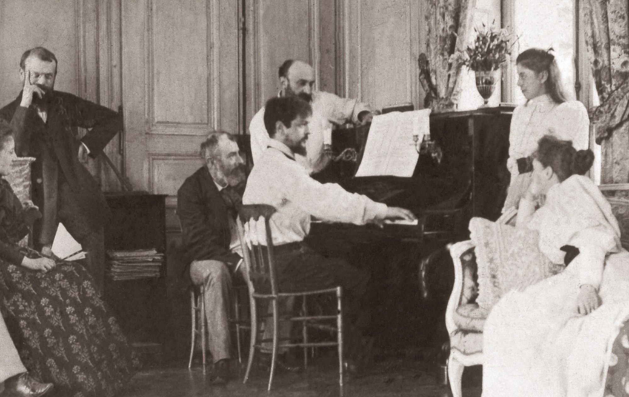 Sepia photo. Debussy playing the piano surrounded by family/friends