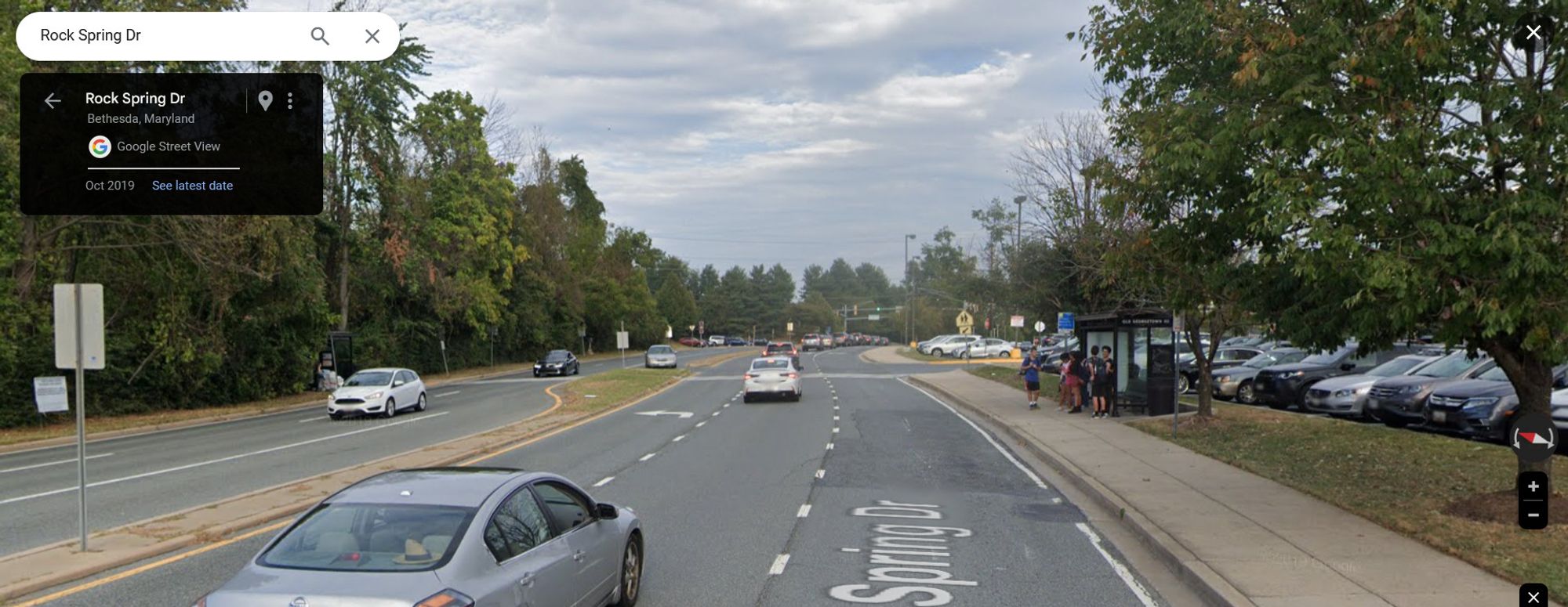 Google Streetview of a six-lane divided road, with a pair of bus stops, and an uncontrolled marked crosswalk between the bus stops. A group of high school students is standing in the bus shelter at one of the bus stops.