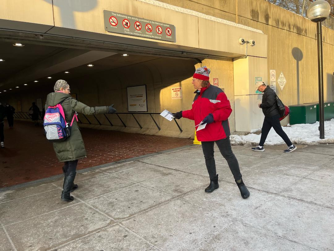 A person bundled up in winter clothing, offering a leaflet to a person also bundled up in winter clothing, on the lower side of the Shady Grove Metro station.