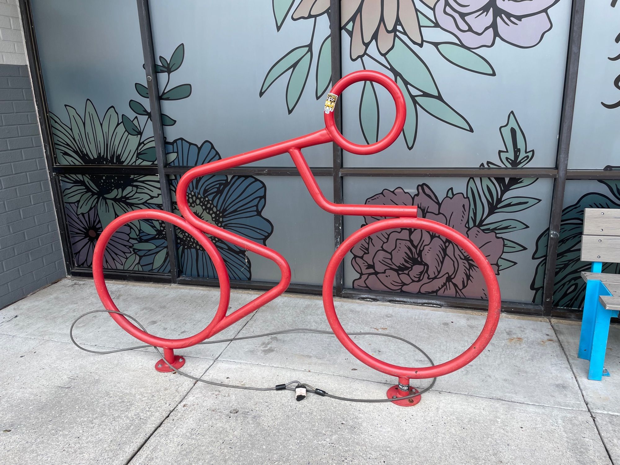 An artistic bike rack outside a grocery store. It's red metal, kind of in the shape of the bike lane stencil only without a helmet. It is raised and bolted to the concrete under the front wheel and back wheel parts.