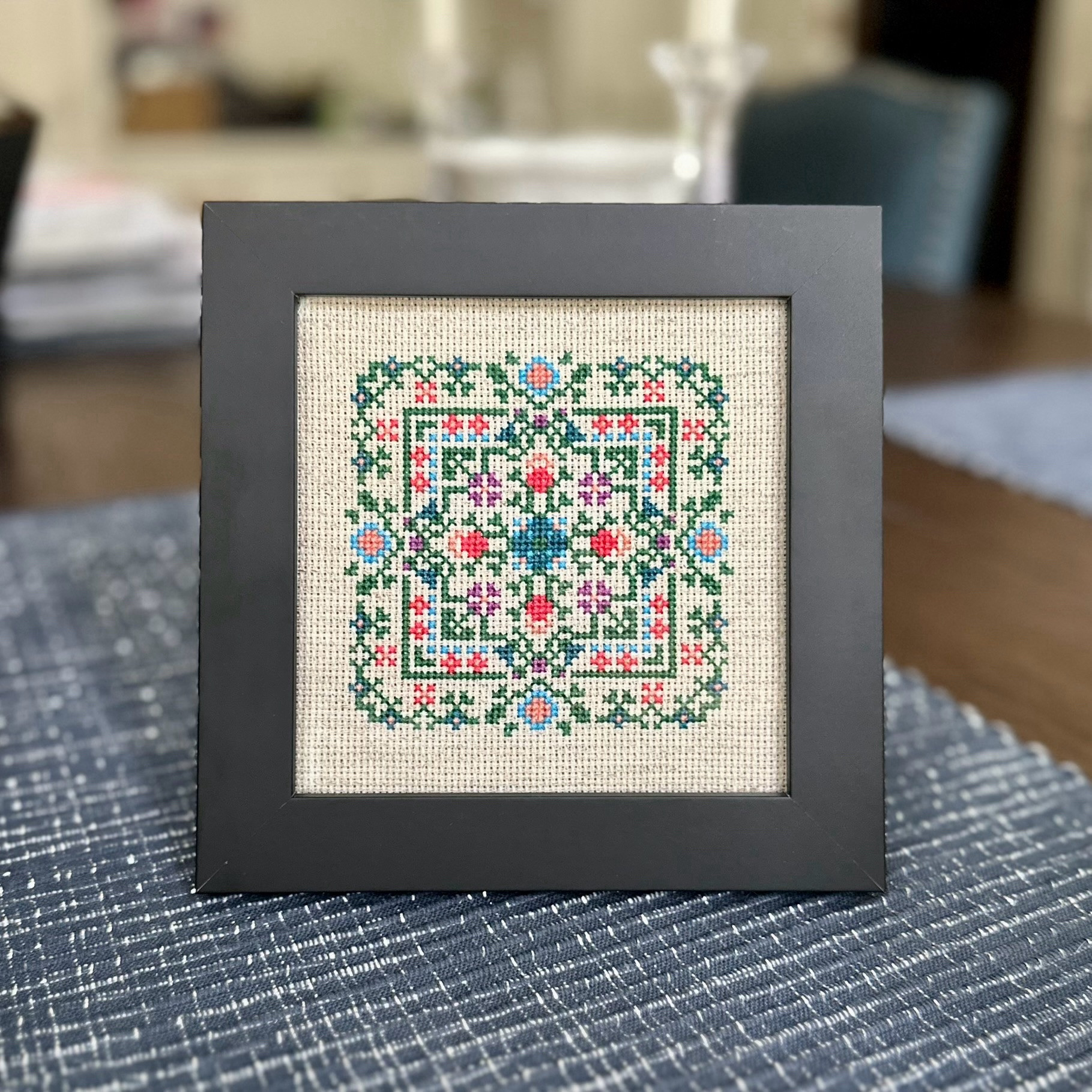 A 4”x4” cross stitch design on natural (beige) Aida in a black 5”x5” frame. The design is symmetric in X and Y, with green leaves around the outside and purple, blue, and coral flowers throughout