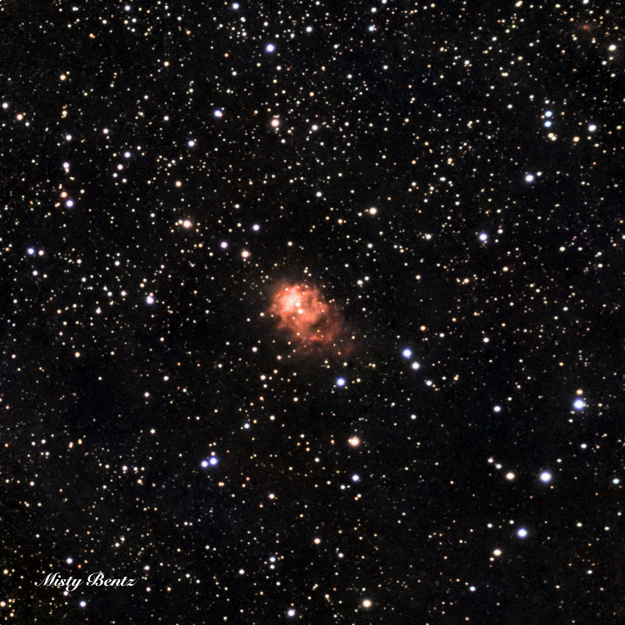 Centered on a black field dotted with stars is a small blue nebula. It is oriented like a number 8 rotated 45 degrees, and is brighter with more defined edges in the top left, while fainter and less defined to the bottom right. Two bright stars are embedded in the middle and upper left of the nebula