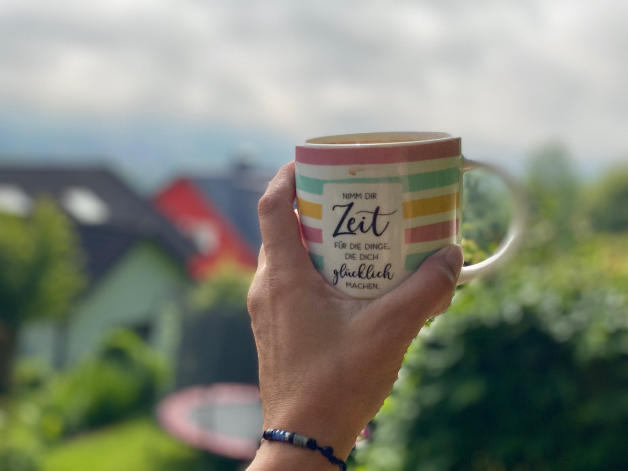 Erster Kaffee an der höchsten Stelle im Garten, weiter Blick. 
Zu sehen meine bunte Kaffeetasse mit dem Spruch „Nimm dir Zeit für die Dinge, die dich glücklich machen“ und meine Hand vor Grünzeug und Trampolin, im Hintergrund verschwommene Häuser.
Die Sonne bahnt sich sanft ihren Weg durch den Morgennebel.