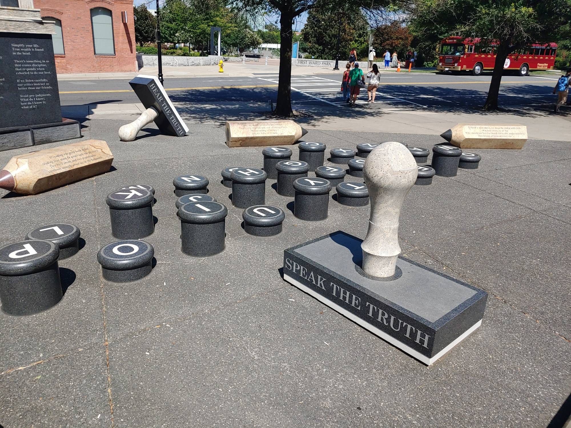 The keys of a typewriter recreated as a sculpture big enough for a person to step from letter to letter, placed across pavement between two sculptures of library stamps (labeled Speak the Truth and Hear the Truth) and three marble pencils (each engraved with a stanza of text) big enough to sit on as benches. A family crosses a crosswalk across the street beyond, headed toward a red bus.