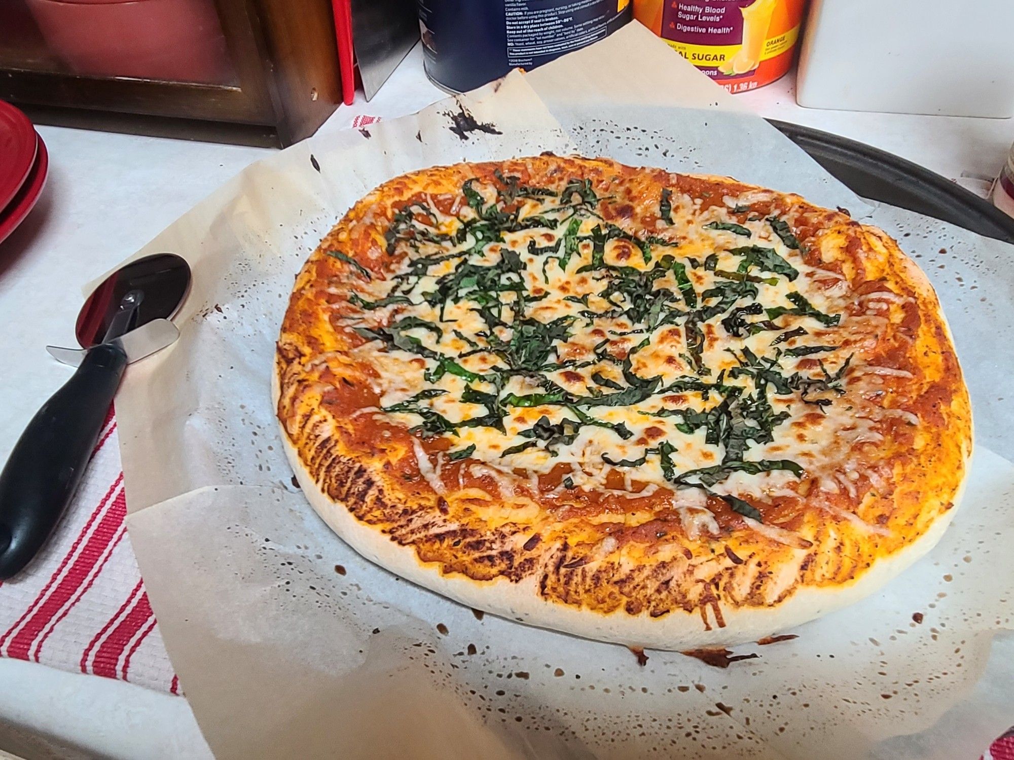 Cheese and basil-topped pizza with stuffed crust, resting on a pizza tray with parchment paper, fresh out of the oven. Pizza cutter for scale on the left.