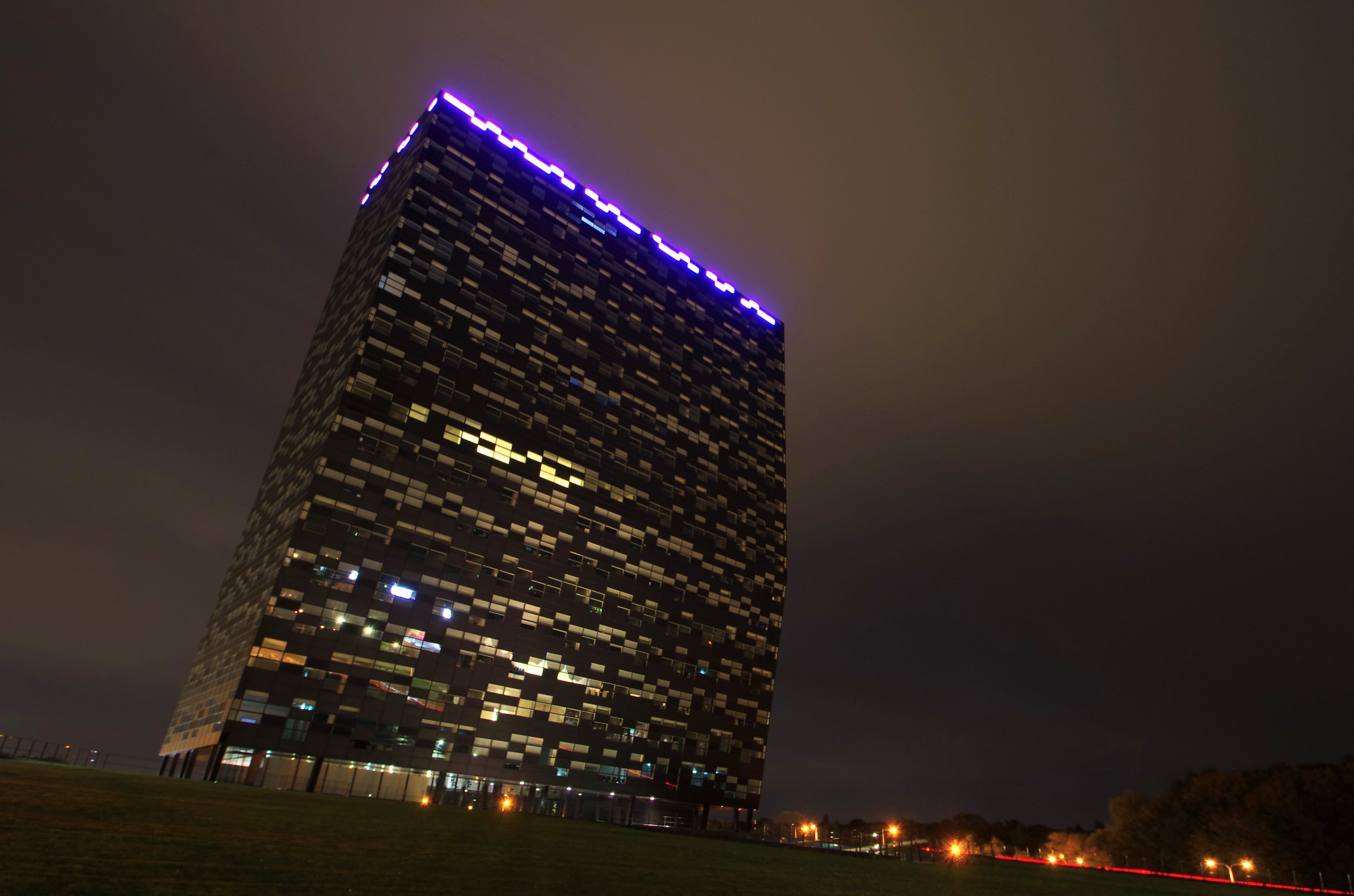 Ein Gebäude in der nacht fotografiert, ober sind lilafarbene Lichter, aus einigen Zimmers scheinen Lichter. Rechts ein roter Lichtstreifen 