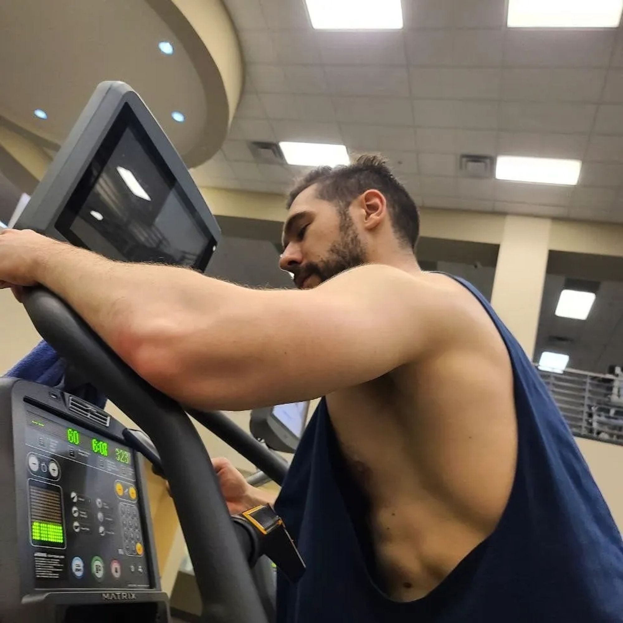 Man sweating on stairmaster, his abs revealed through side of overly large tanktop