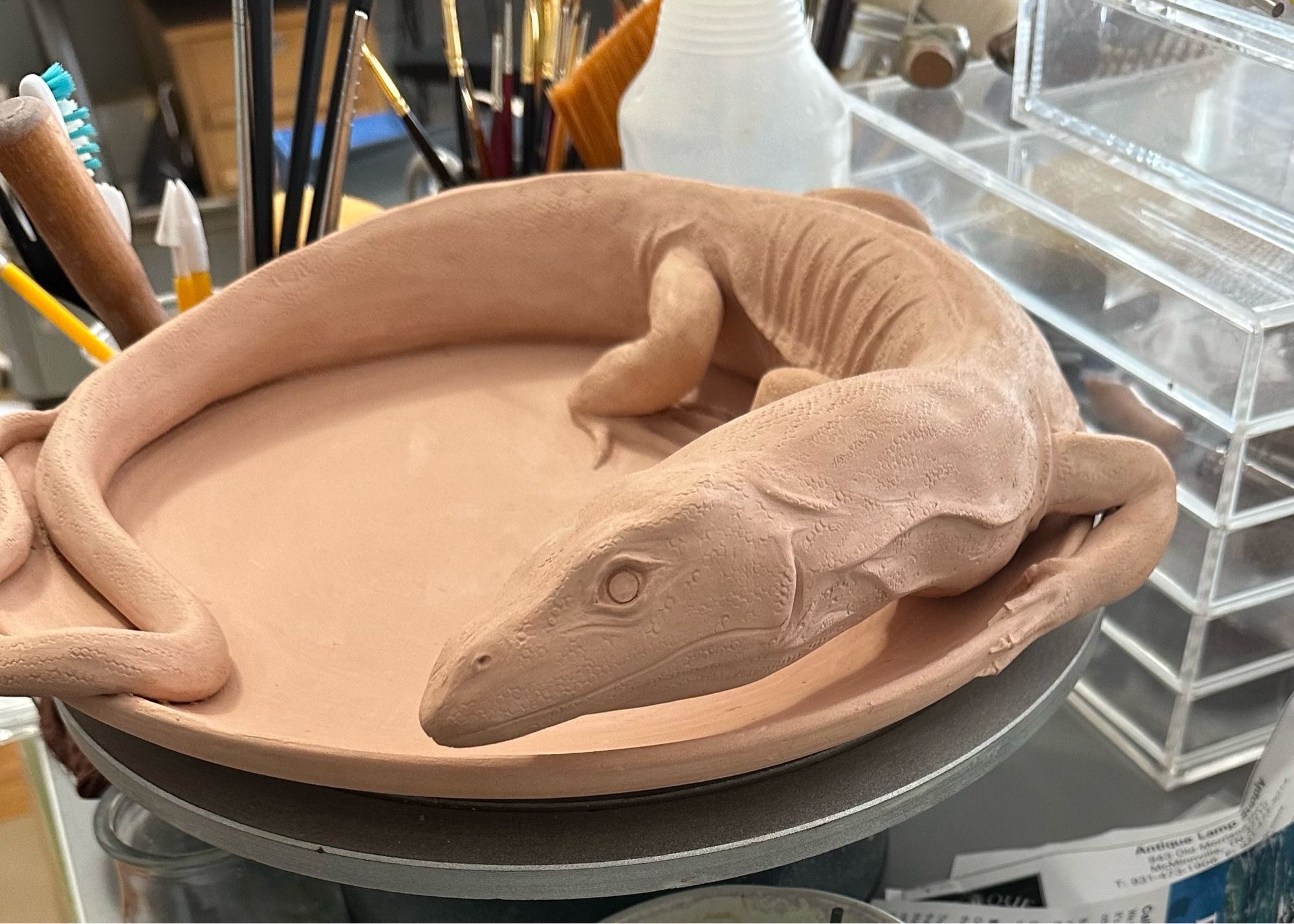 A bisque fired ceramic monitor lizard curled around the edge of a plate is giving the camera skeptical side eye.