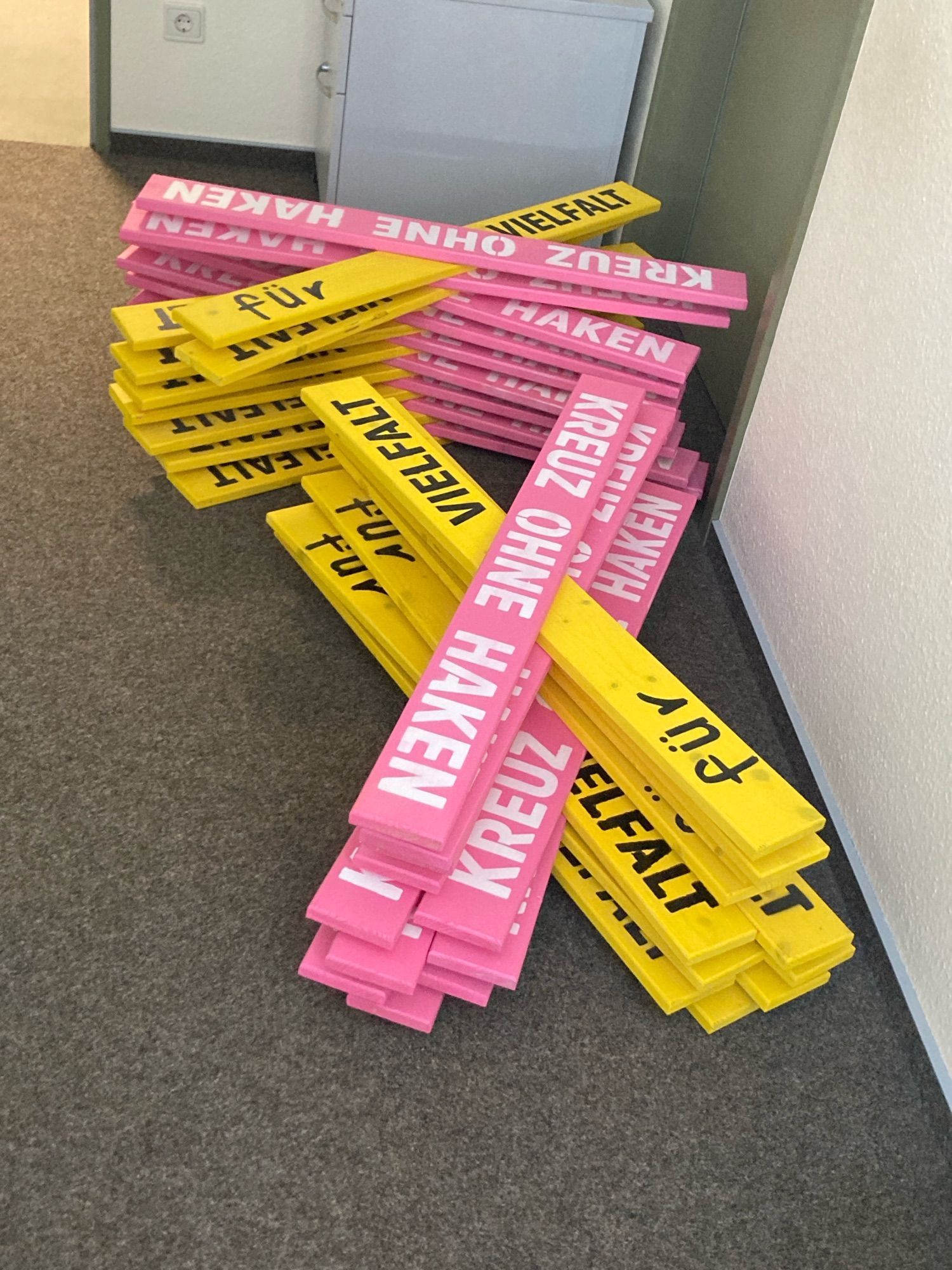 Pink-gelbe Holzkreuze liegen auf einem Büroteppichboden. Sie tragen auf dem pinken Arm die Aufschrift „Kreuz ohne Haken“ in weißen Großbuchstaben und auf dem gelben Arm „für Vielfalt“ in einer schwarzen Mischtypo aus großen und kleinen Buchstaben.