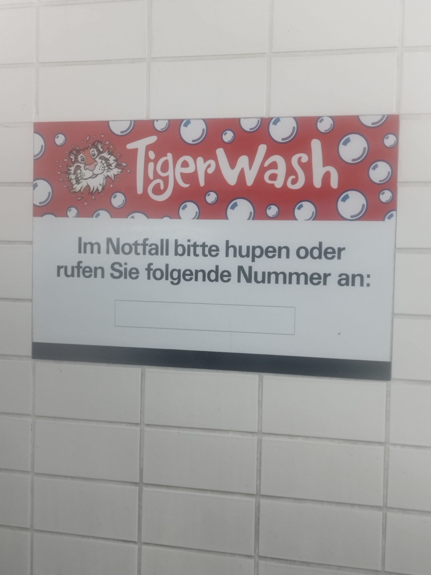 Schild in einer Waschstraße auf einer gefliesten Wand. 
Oben das Logo "Tigerwash"
Text: Im Notfall bitte hupen oder rufen Sie folgende Nummer an: