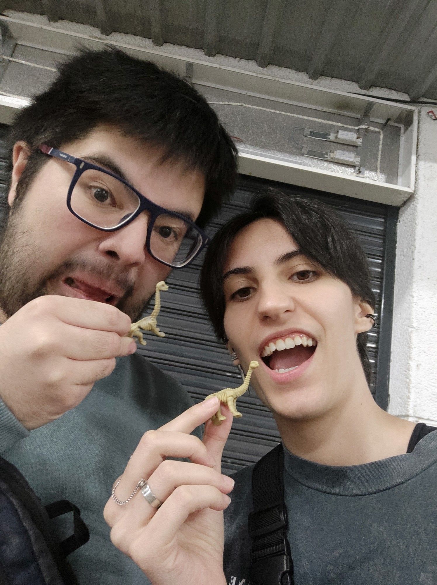 Una foto de mi amigo Gaby y yo. Salimos con la boca abierta intentando poner cara amenazante, imitando a un dinosaurio (?). Ambos sostenemos una figurita de un esqueleto de dinosaurio, Gaby (a la izq) un brontosaurio y yo (dcha) un diplodocus. Los dos llevamos una camiseta de color gris/negro verdoso. Gaby lleva gafas, tiene barba y el pelo negro. Yo llevo algunos pendientes y piercings en la oreja y soy morena en general también