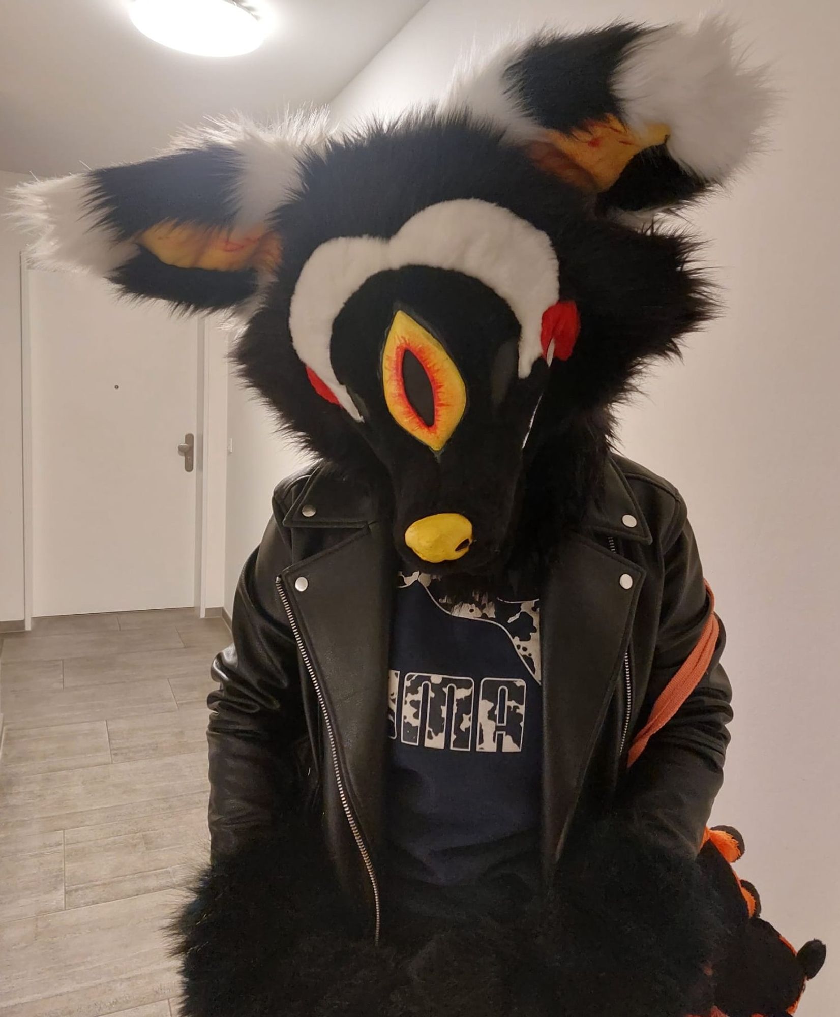 Cyclops fox fursuit with black and white striped ears, a yellow eye with veins, and red circels on it checks, with a sly expression