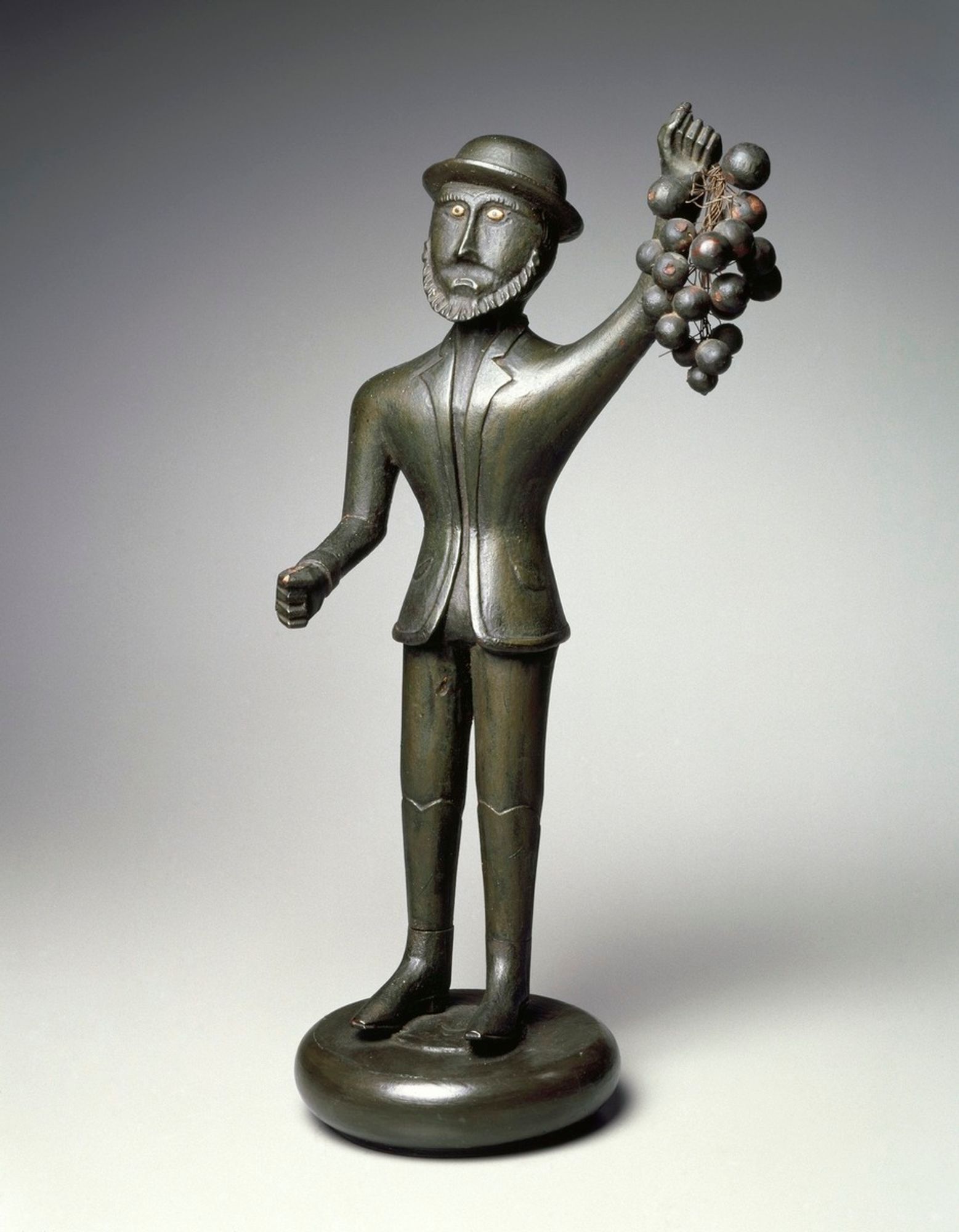 Carved wooden figure of man, primitive style, painted gray-green.  Man stands on a low cylindrical base and holds aloft an oversized bunch of grapes in proper right arm; his proper left arm is bent at elbow and extends perpendicular to the figure's body.  He is dressed in a suit, knee-high boots, and Derby hat.  Eyes are bone.  Grapes are attached to wires threaded through hole in figure's hand.  Left hand is in a gripping position with hole bored through the fist, probably for attaching a now lost component (perhaps a wineglass).

Condition: Very good; minor wear, appears to be missing a component.
