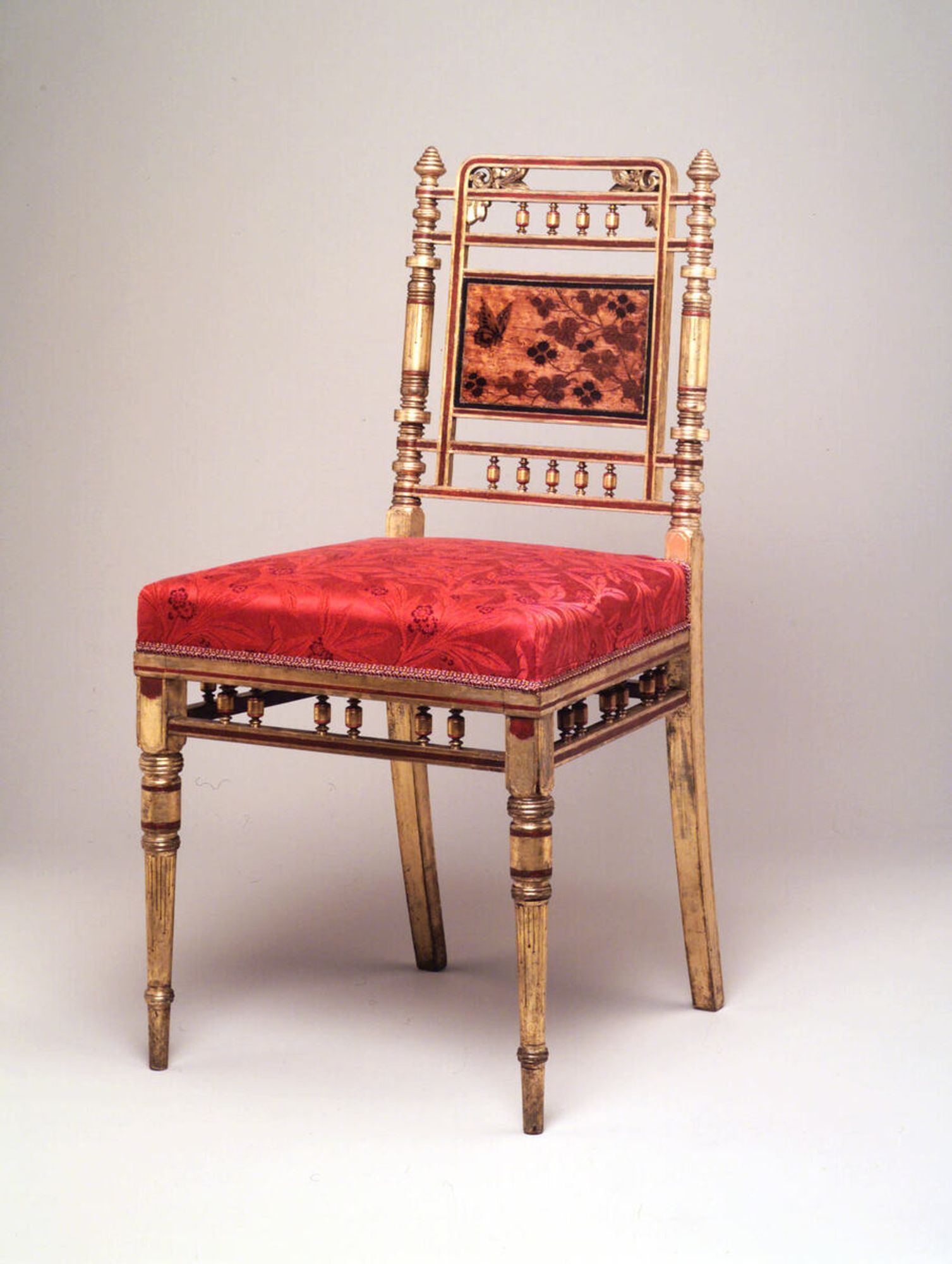 Side Chair, gilded maple,bird's-eye maple, mahogany and rosewood (marquetry), ash (secondary wood), red and black paint, modern upholstery. Delicately proportioned turned, incised, carved and inlaid chair in the Anglo- Japanese taste. Simple square-profile upholstered seat. Gilded square-profile rear legs splayed backward rise to intricately turned back stiles highlighted with red paint capped with graduated conical finials. Stiles connected by two pairs of narrow square-profile horizontal braces above seat and at crest. Five short turned spindles connect lower braces and four connect upper pair. Rectangular bird's-eye maple marquery panel, framed by red and black painted borders, at center of back supported by square-profile vertical braces that rise from lower cross brace and intersect upper pair of braces and that curve to form flat crest rail with symmetrical carved and cut-out leafy motifs inside each upper curved corner. Panel decorated with asymmetrical Japanese-style floral decoration: bifurcated flowering and leafy branch rises from proper left lower corner of panel with butterfly at proper right upper corner. Turned and incised downward tapering front legs highlighted with red paint rise to square blocks below seat rail. Narrow square-profile horizontal stretchers below seat rails connected to seat rails by three pairs of short turned spindles on sides and front.

CONDITION - Overall losses to gilding and painted areas. Marquetry panel on back upholstered over; tack holes now around perimeter. Surface conserved by Pascale Patrice and seat conserved by Nancy Britton, both from the Metropolitan Museum of Art (summer/Fall 2000) . See cosevation reports for frame and upholstery in file.