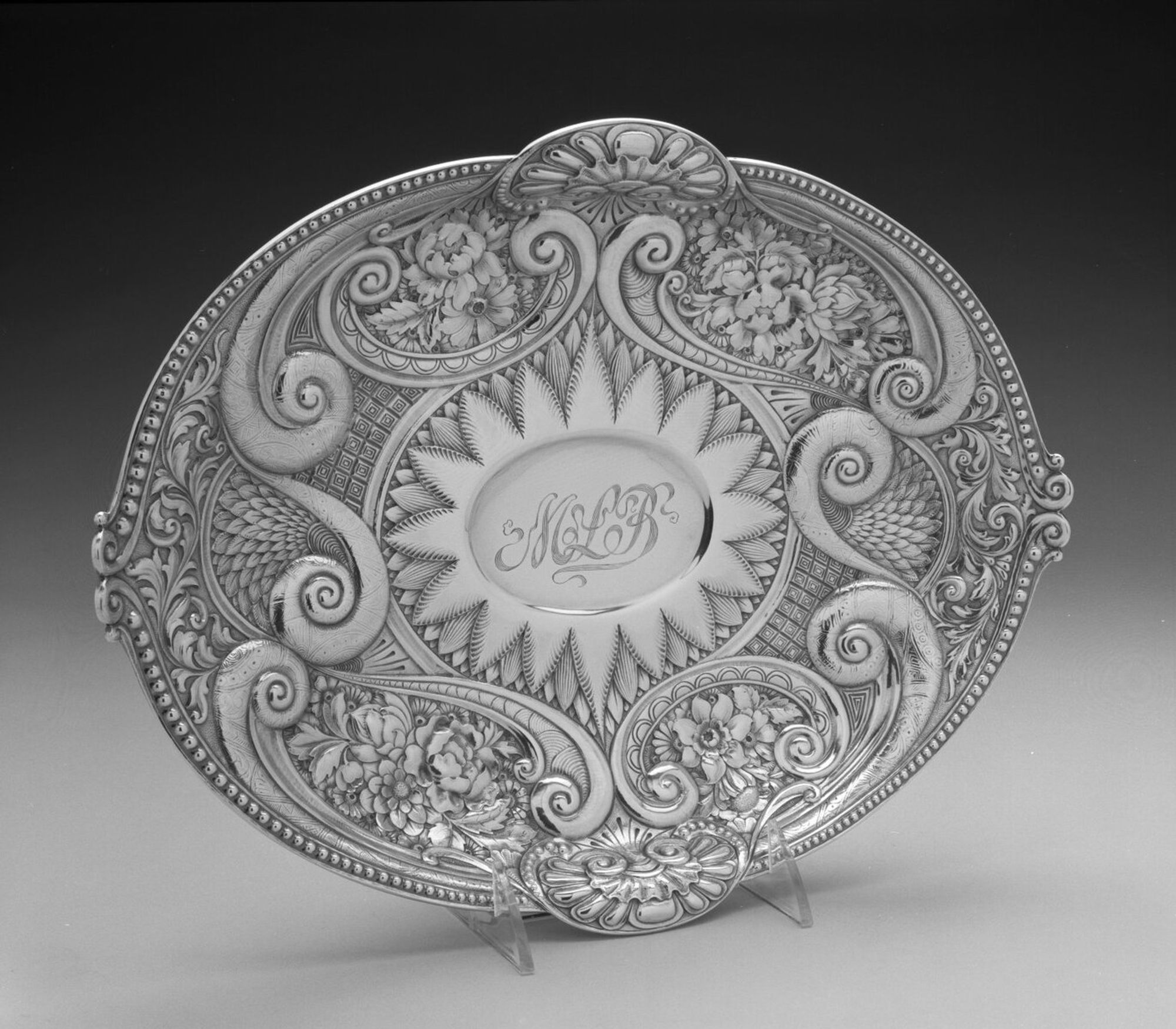 Footed Dish: silver. Oval concave dish rests on a plain oval ogee foot. The dish is elaborately repoussed around a central oval void containing the initials in script: MLB. Surrounding this is an area of layered pointed petal forms. Followed by an area of broad volutes, most engraved with dense abstract pattern. The volutes rest on a diaper ground. At each end of the dish, volutes surround layered feathers with scrolling acanthus foliage above. At each long side, volutes surround areas of densely packed blossoms. The dish has a beaded rim broken on each long side by a c-shaped cartouche, containing abstract shell forms, rising above the rim, and terminating on each short side with small opposed c-scrolls.


CONDITION - Fine.