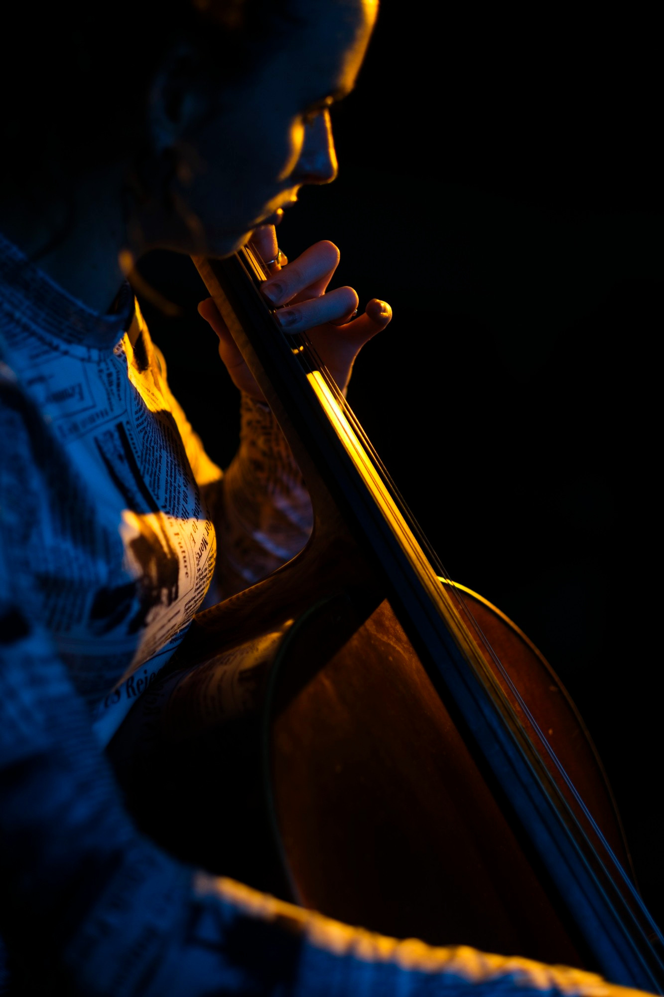 The image shows a person playing a cello. The lighting is dramatic, highlighting only parts of the person and the cello, creating a high-contrast effect. The person’s face is partially visible as they look down at the instrument, with their hands positioned as if they are playing it. The background is dark, emphasizing the subject and adding to the moody atmosphere of the photograph.