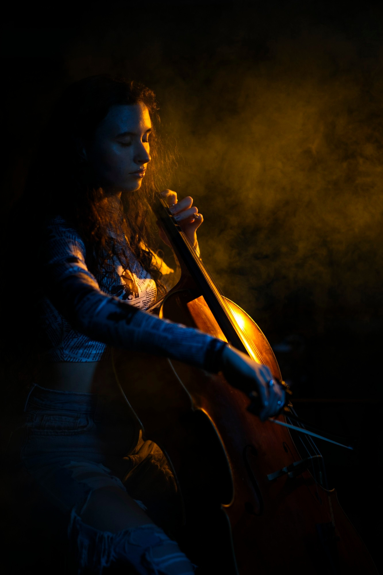 The image shows a person playing a cello in a dimly lit setting. A strong light source highlights the dust or rosin particles in the air around the bow and strings, creating a dramatic effect.