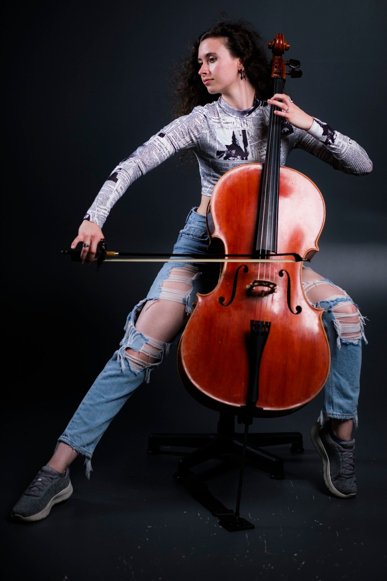 The photo shows an individual playing a cello. They are seated and appear to be in the act of playing the instrument. The individual is wearing a patterned long-sleeve top, ripped blue jeans, and grey sneakers. The cello has a warm wood finish and is supported by an endpin stand.