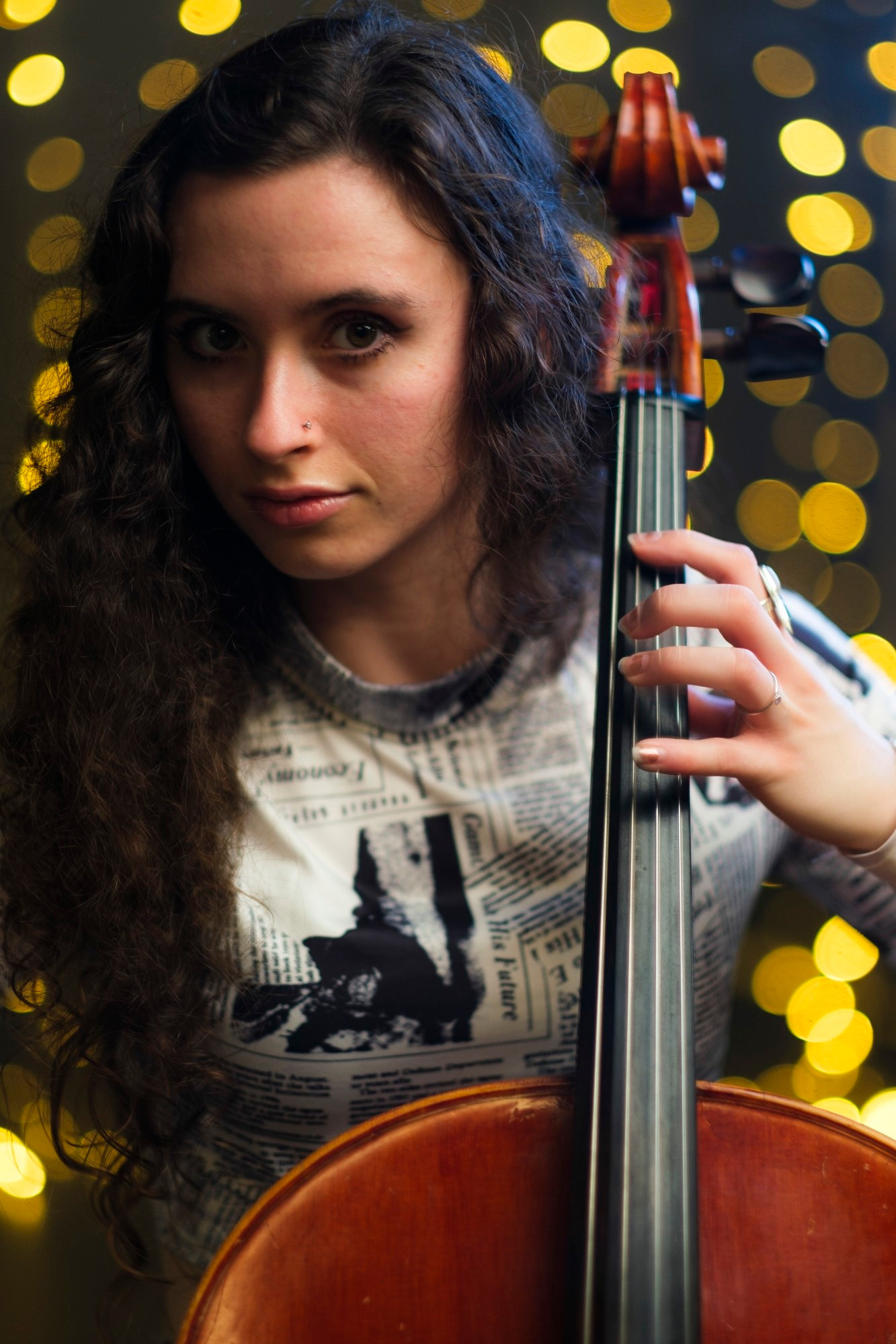 The photo shows an individual holding a cello. The person is seated and wearing a t-shirt with a newspaper-style print that includes images and text. The background is blurred with a bokeh effect, featuring numerous small, circular light spots, suggesting the photo was taken in a low-light environment with distant light sources.