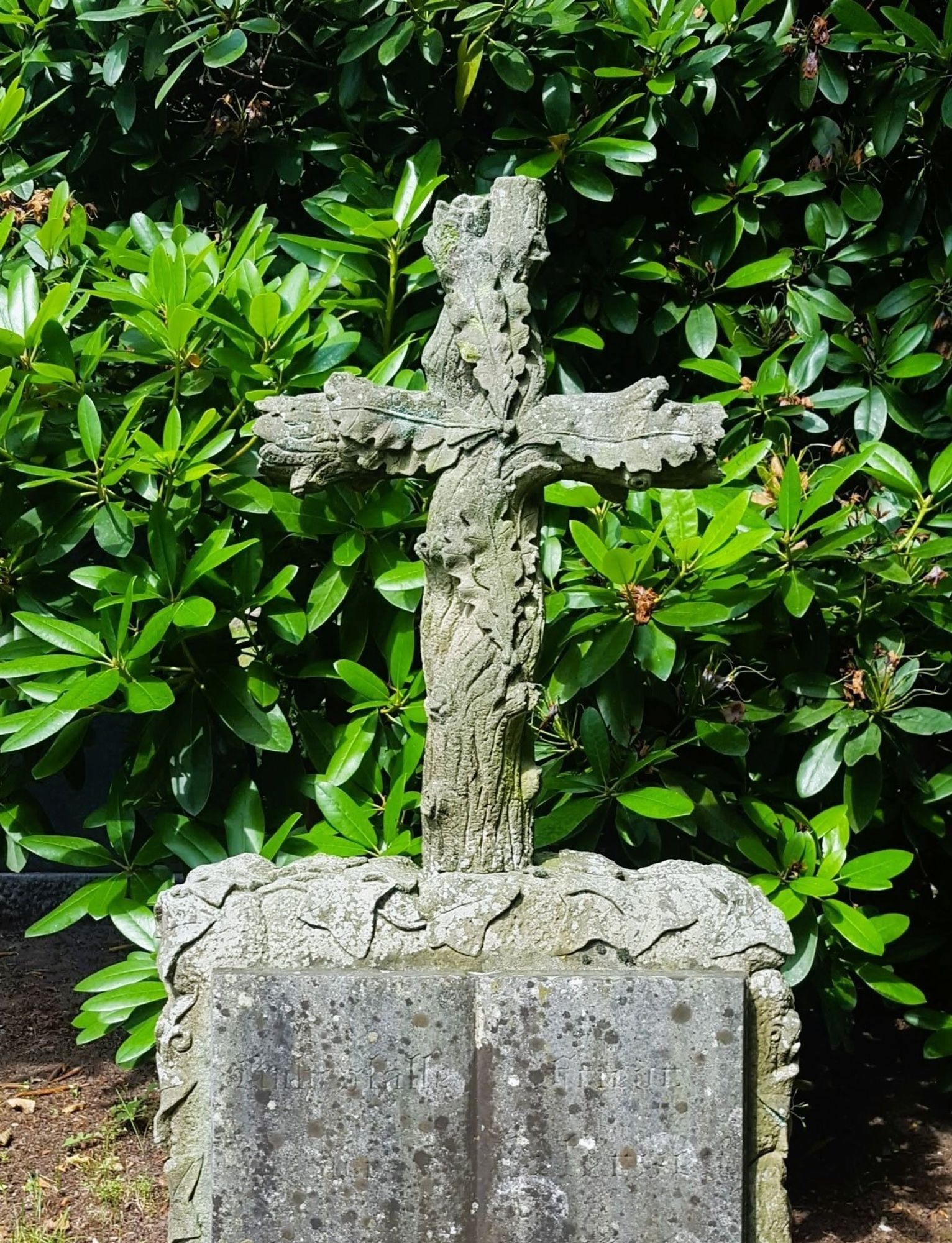 ein Steinblock mit Inschrift, darauf ein Steinkreuz mit der Gestalt eines Eichenstammes und Eichenblättern
a stone cross, that looks like an oak trunk with leaves