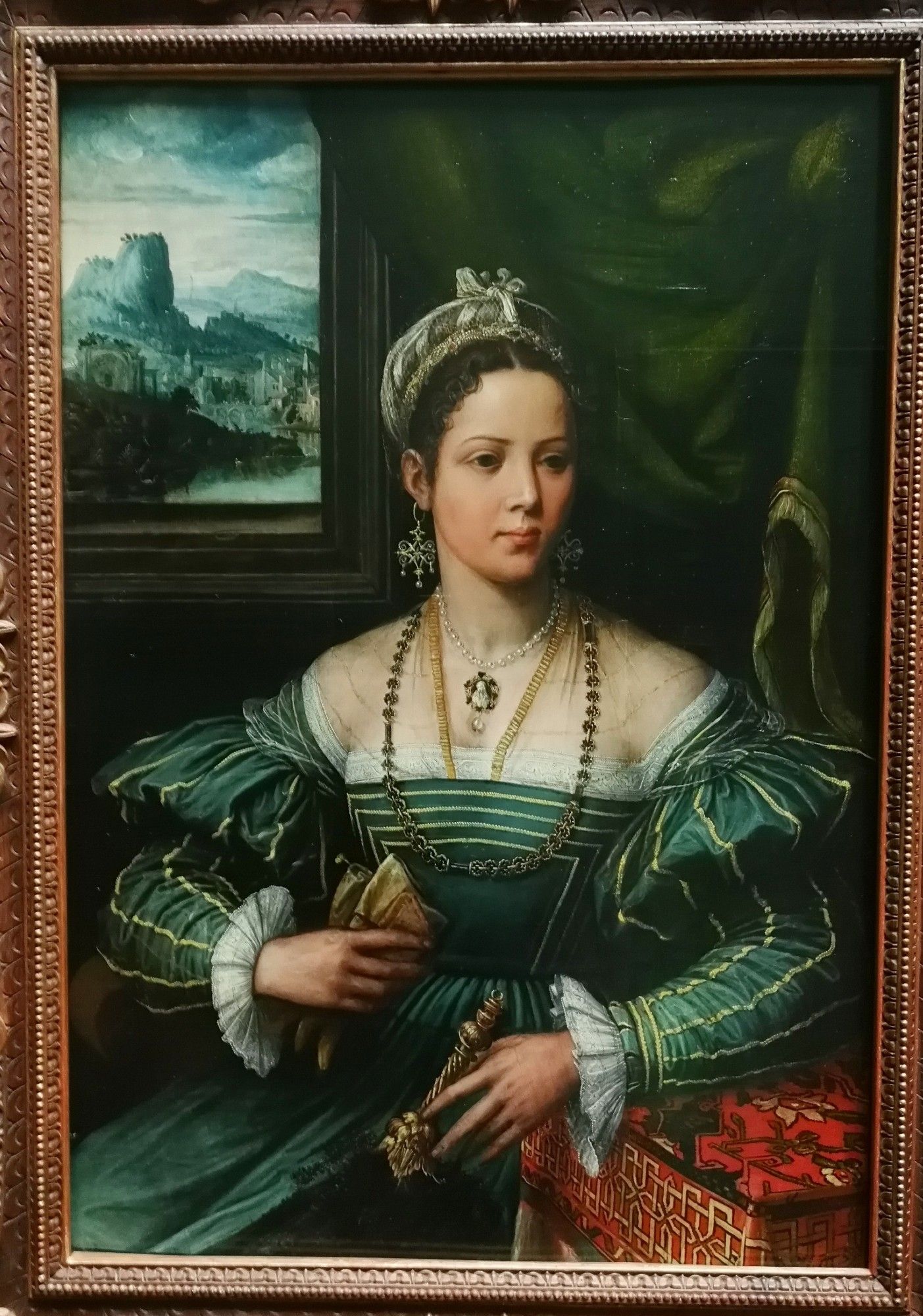 Eine Dame in einem grünen Kleid und mit viel Schmuck sitzt vor einem grünen Hintergrund, an der Wand hängt ein Gemälde mit Landschaftsdarstellung