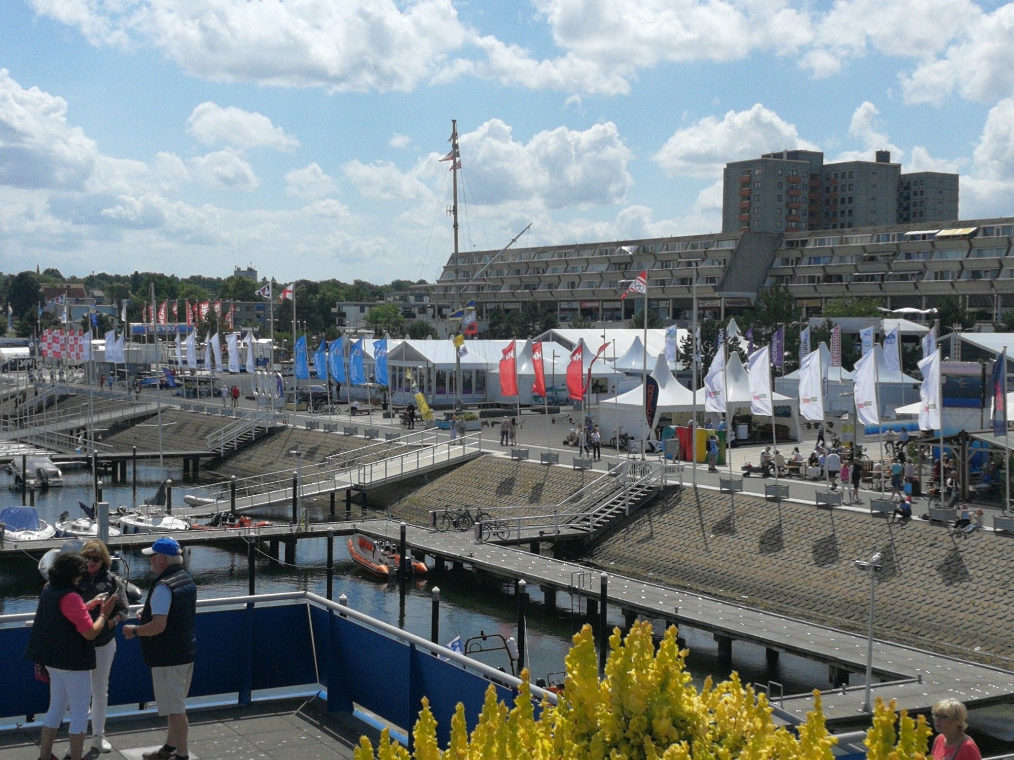 Blick vom Aussichtsturm zur Uferpromenade - auf dieser stehen Zelte mit Gastronomie, der Weg ist mit Fahnen geschmückt, im Hintergrund eine Wohnanlage