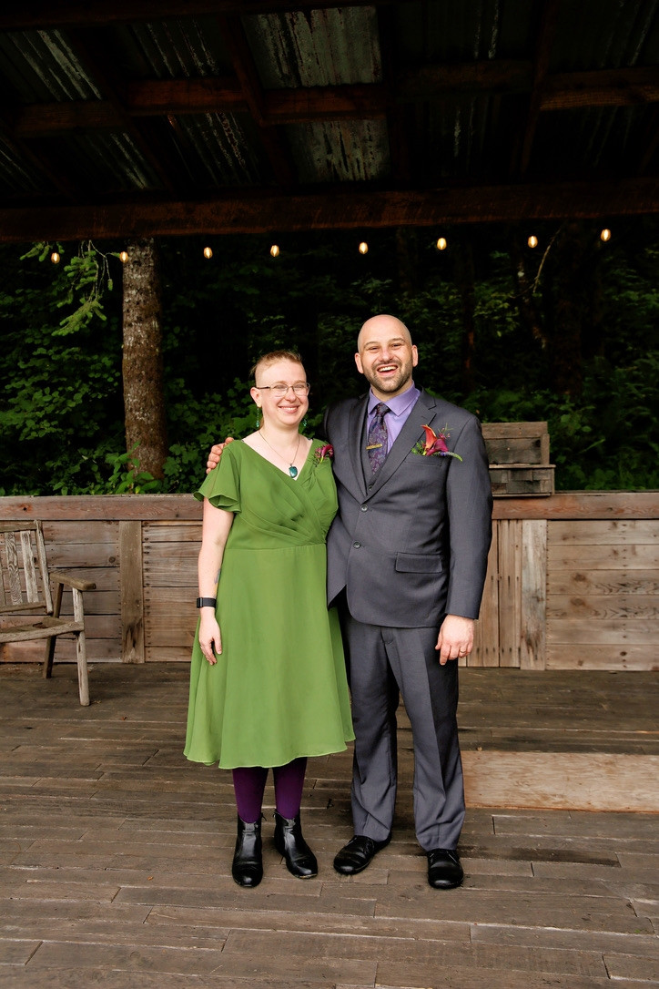 Aradia and Seth side by side in their wedding best. Aradia is dressed in a green dress as his groomsmaid, Seth is dressed in a nice suit with a purple tie and shirt. They are standing on an outdoor wooden stage and are both grinning in the aftermath of Seth becoming a happily married man. They are both white, he is slightly taller and has significantly less hair.