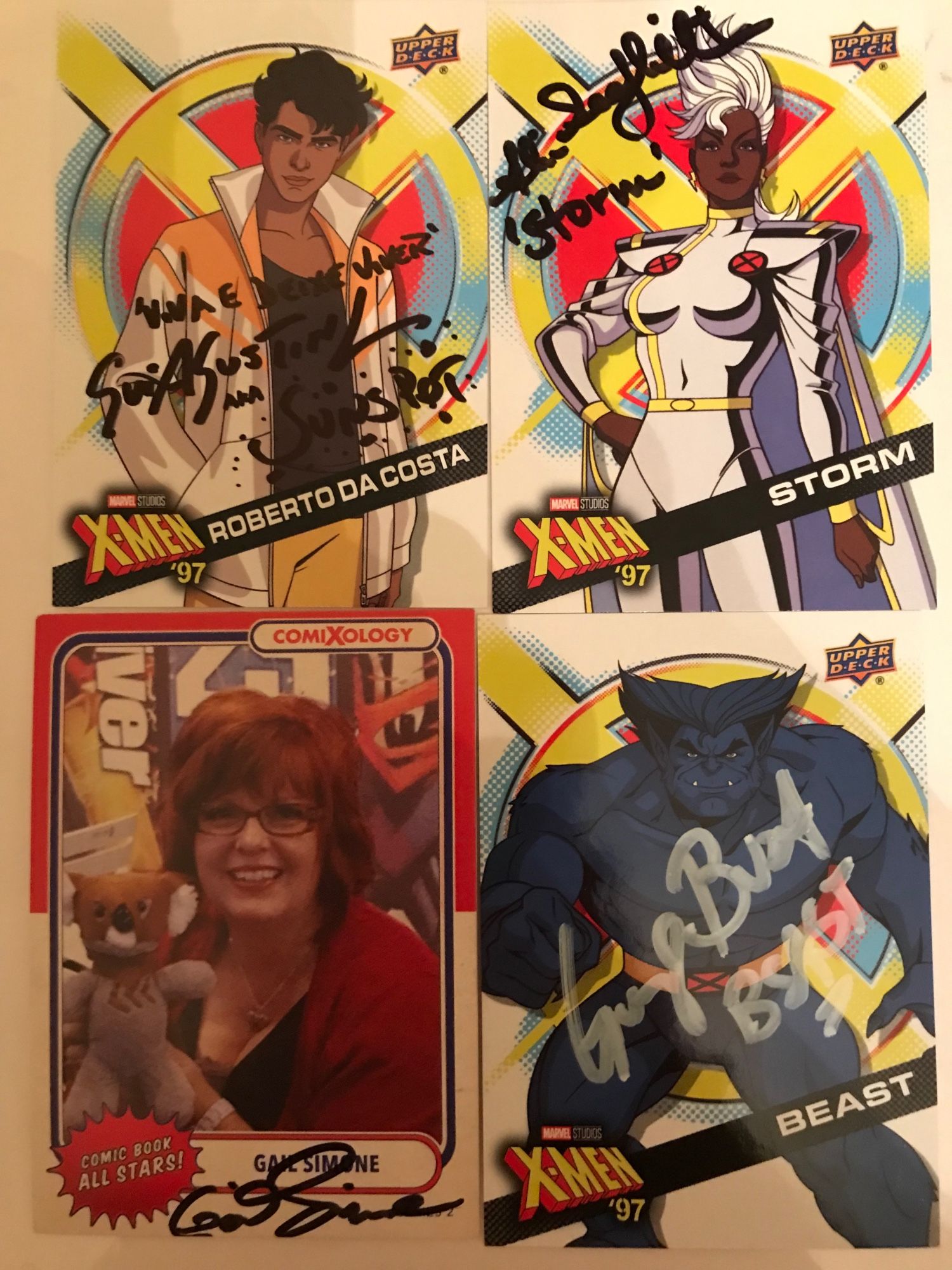 Top left card says X-Men ‘97: Roberto Da Costa by Upper Deck, signed by Gui Agustini, who signed, “Viva e deixe viver,” which is Spanish for, “Live and let live,” his name, and then “Sunspot.”

Too right card says X-Men ‘97: Storm by Upper Deck, signed by Alison Sealy-Smith, “Storm.”

Lower left card says Comic Book All Stars! by comiXology, Gail Simone, signed by Gail Simone.

Lower right card says X-Men ‘97: Beast by Upper Deck, signed by George Buza, “Beast.”