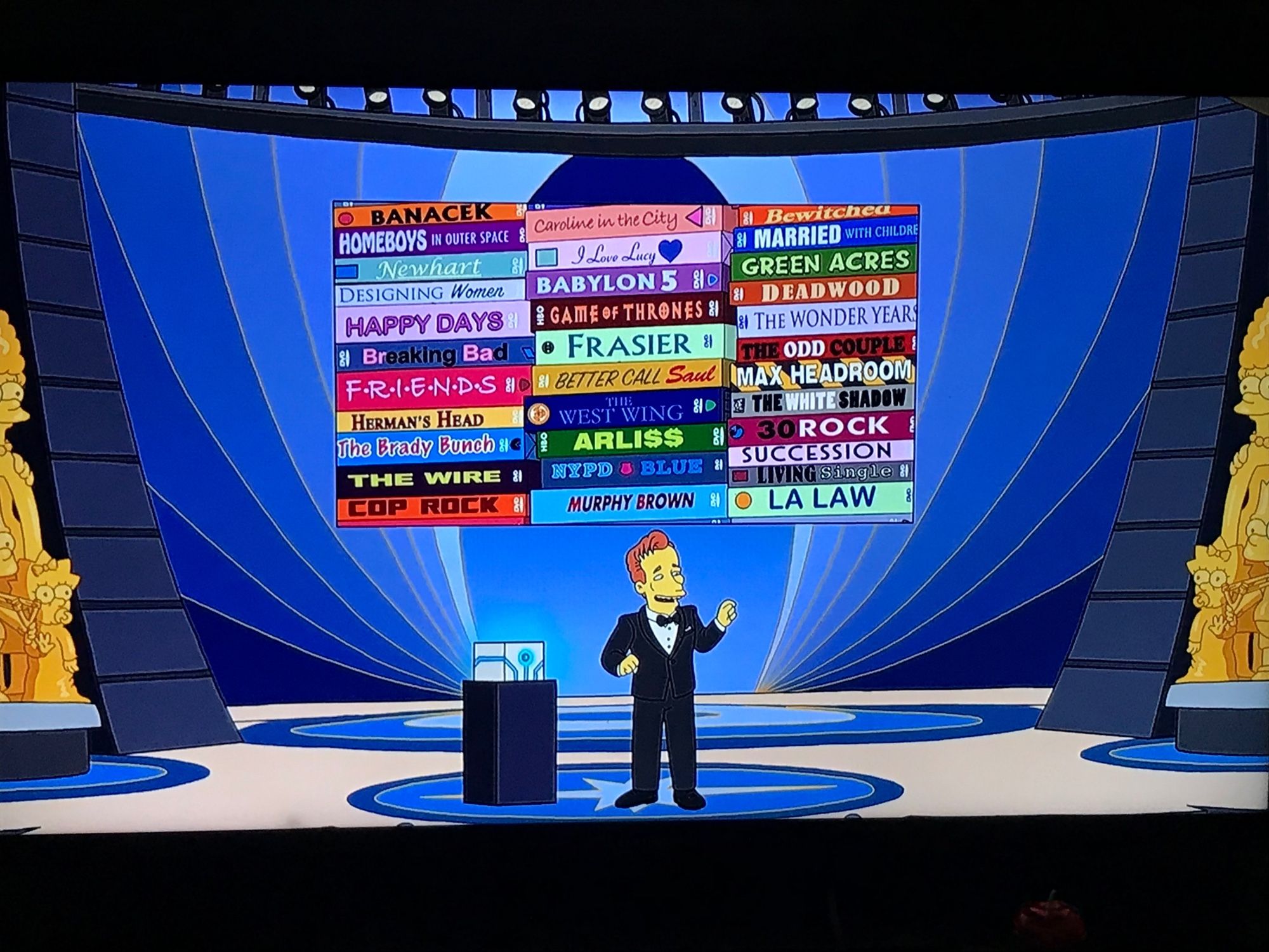 Screenshot from The Simpsons season 36 episode 1, “Bart’s Birthday,” over Conan O’Brien’d head, the TV shows listed by column.

In column 1 from top to bottom, Banacek, Homeboys in Outer Space, Newhart, Designing Women, Happy Days, Breaking Bad, Friends, Herman’s Head, The Brady Bunch, The Wire, and Cop Rock.

In column 2 from top to bottom, Caroline in the City, I Love Lucy, Babylon 5, Game of Thrones, Frasier, Better Call Saul, The West Wing, Arli$$, NYPD Blue, and Murphy Brown.

In column 3 from top to bottom, Bewitched, Married with Children, Green Acres, Deadwood, The Wonder Years, The Odd Couple, Max Headroom, The White Shadow, 30 Rock, Succession, Living Single, and LA Law.