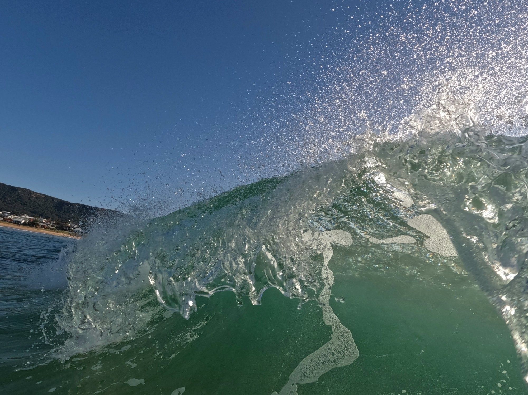 Photo of a wave breaking, early morning. Curve of green. Gleam of spray against a blue sky. Glimpse of sandy beach & escarpment. Bulli, NSW.