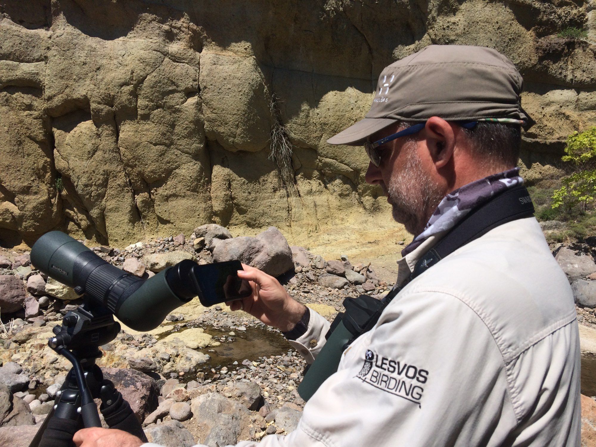 The poster wearing a beige shirt (Lesvos Birding logo on the arm featuring a stylised Little Owl), black and white cammo neck scarf (buff) and khaki green peaked hat with a pair of green binoculars around his neck on a wide, black neck strap. I am using my iPhone with an adaptor on my tripod-mounted spotting scope to film Crag Martin in Lesvos, Greece.