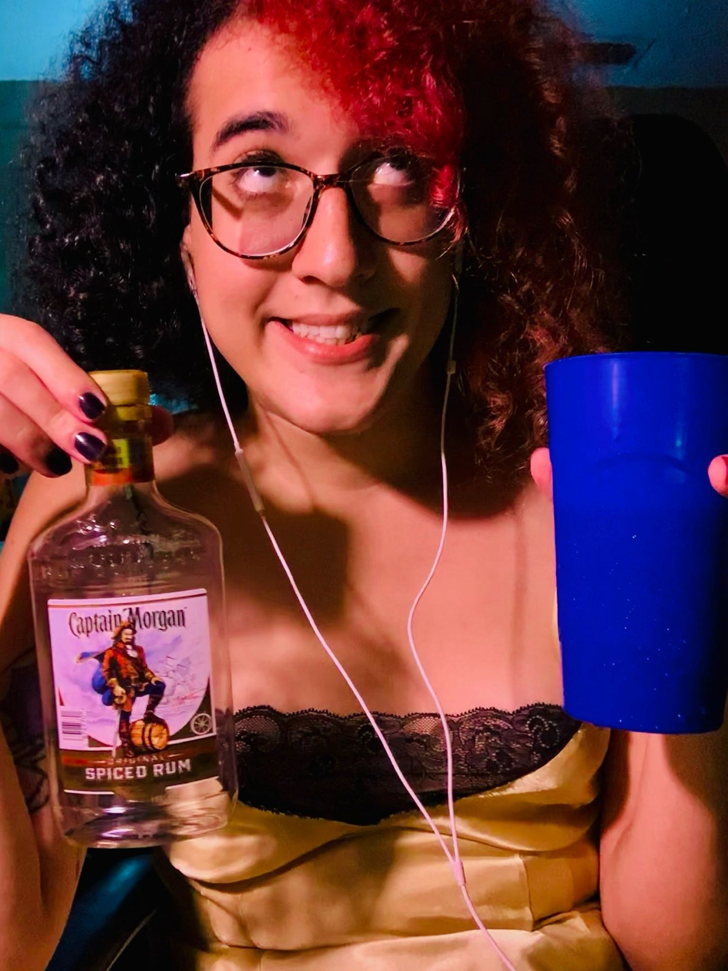 vivi in 2020, with much worse hair, looking very intoxicated holding an empty rum bottle and a tall blue cup that is definitely filled with rum. she’s wearing a gold satin nightgown that i actually really miss being able to fit into