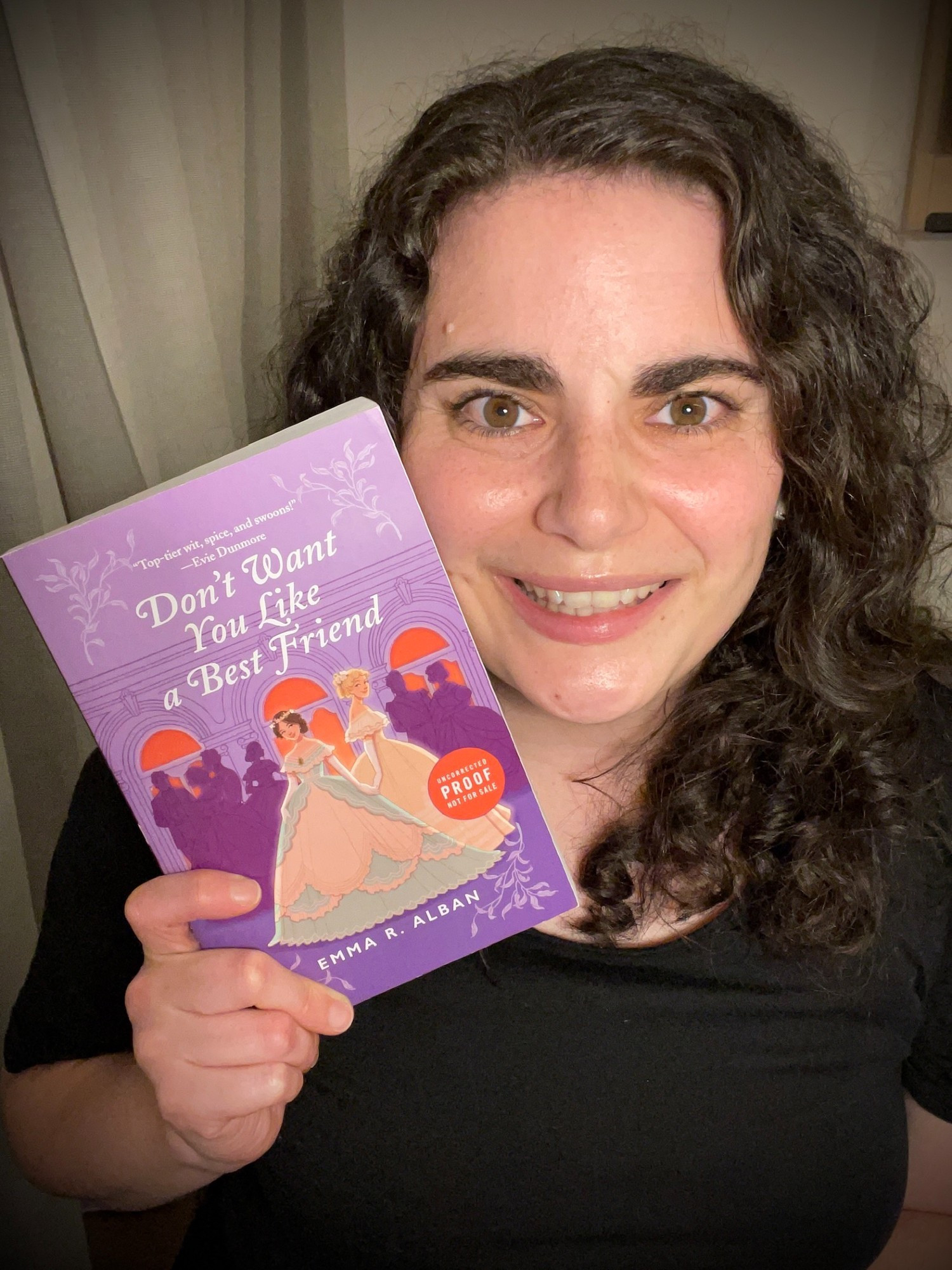 A woman with curly brown hair, brown eyes, and dark eyebrows, wearing a black tee shirt holds the uncorrected proof copy of "Don't Want You Like a Best Friend": Purple cover, with one brunette Victorian debutante in green, holding the hand of a blond Victorian debutante in yellow, in front of purple silhouetted Victorian partygoers, all in front of coral windows in a ballroom. Author: Emma R. Alban