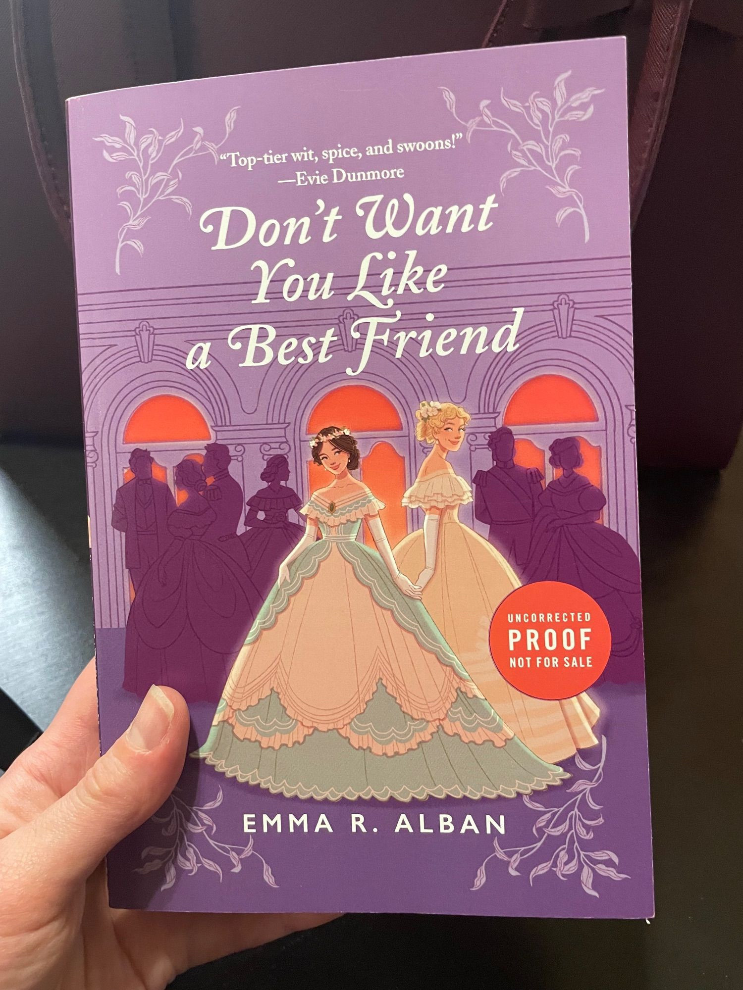 A hand holding the uncorrected paperback proof of debut novel DON'T WANT YOU LIKE A BEST FRIEND: Two Victorian debutantes, one brunette in a green and creme dress, one taller and blonde in a gold dress, stand slyly holding hands in front of a silhouetted purple crowd of partygoers, in front of deep coral windows. Author: Emma R. Alban. Blurb at top of page: "Top-tier wit, spice, and swoons!" - Evie Dunmore.