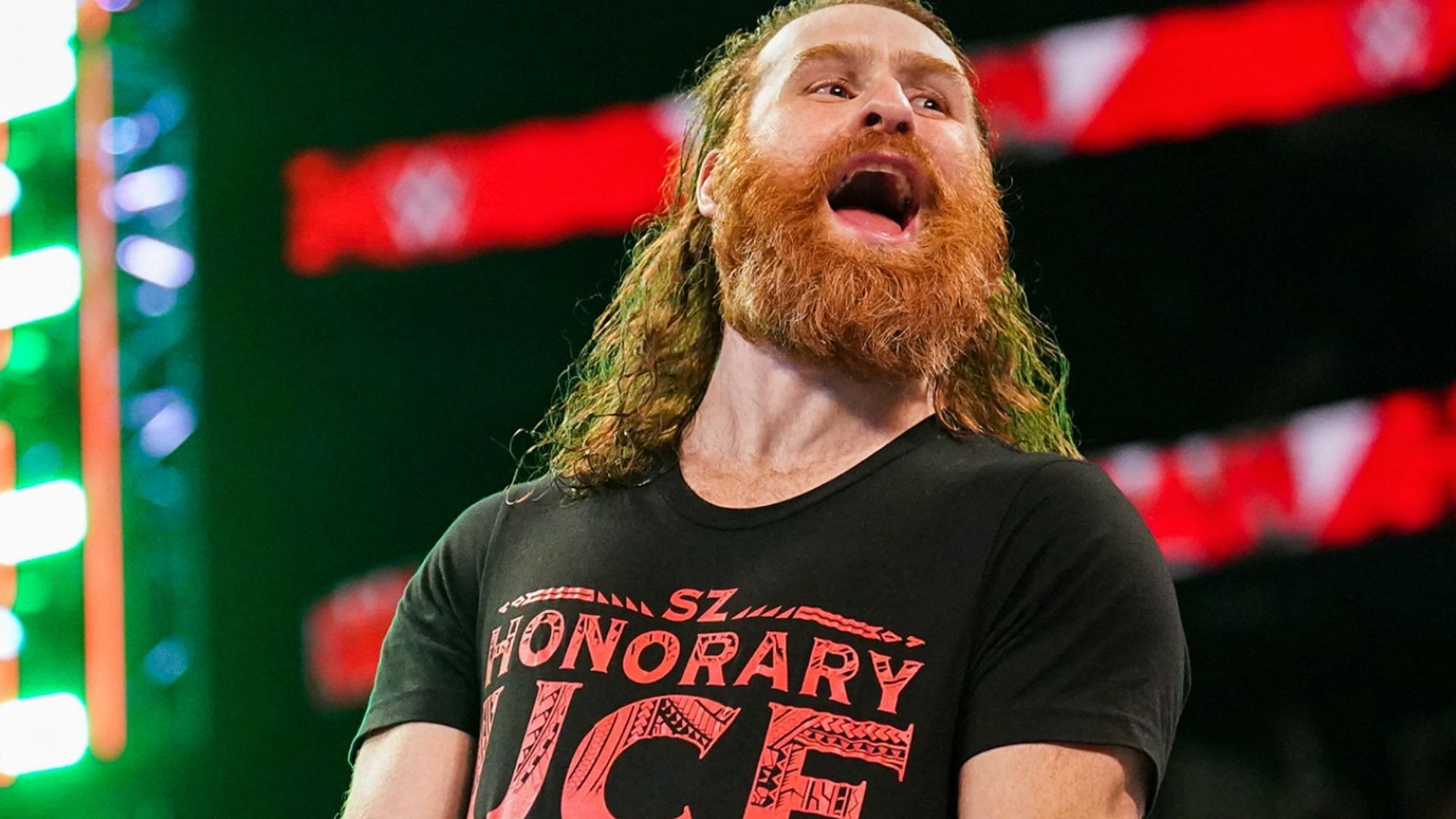 Sami Zayn, a pro wrestler, who has long ginger hair & a prominent beard. He's standing in the ring wearing a shirt that says "SZ - Honorary Uce"