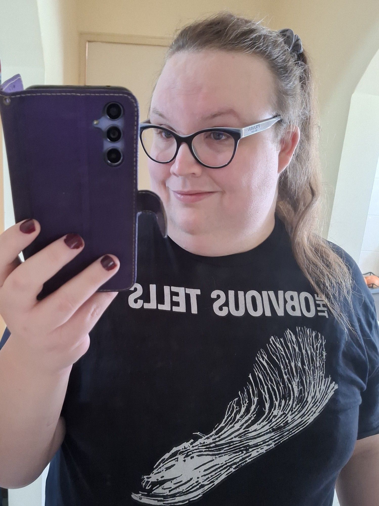 Me taking a mirror selfie. I'm a white genderqueer person with long dirty blonde hair tied up in a high ponytail. I've got glasses & am wearing an Obvious Tells t-shirt