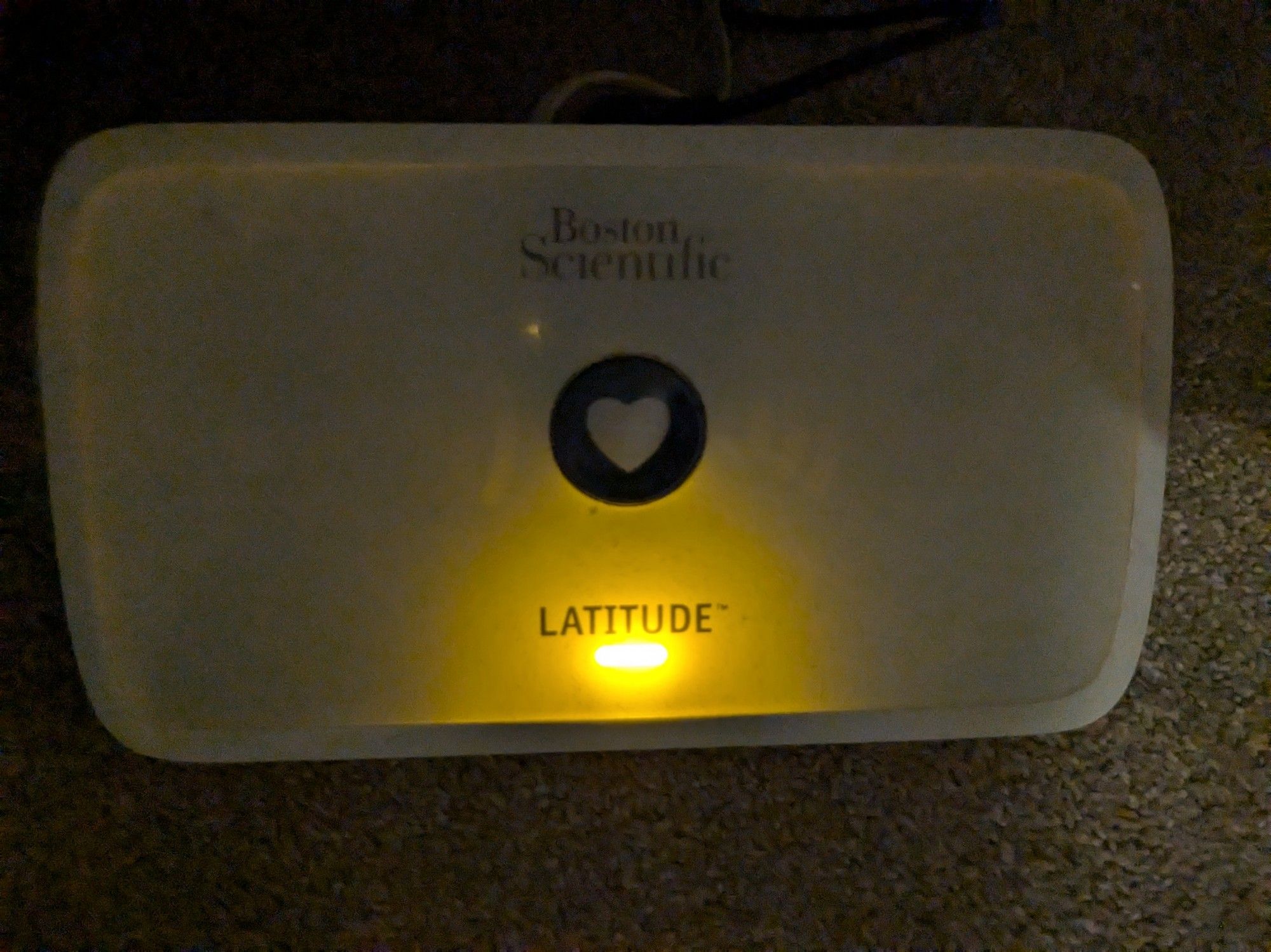 Picture of ICD remote monitor, a white plastic box with a heart button in the middle. Reads Boston Scientific and Latitude. There is a yellow light at the bottom of the box.