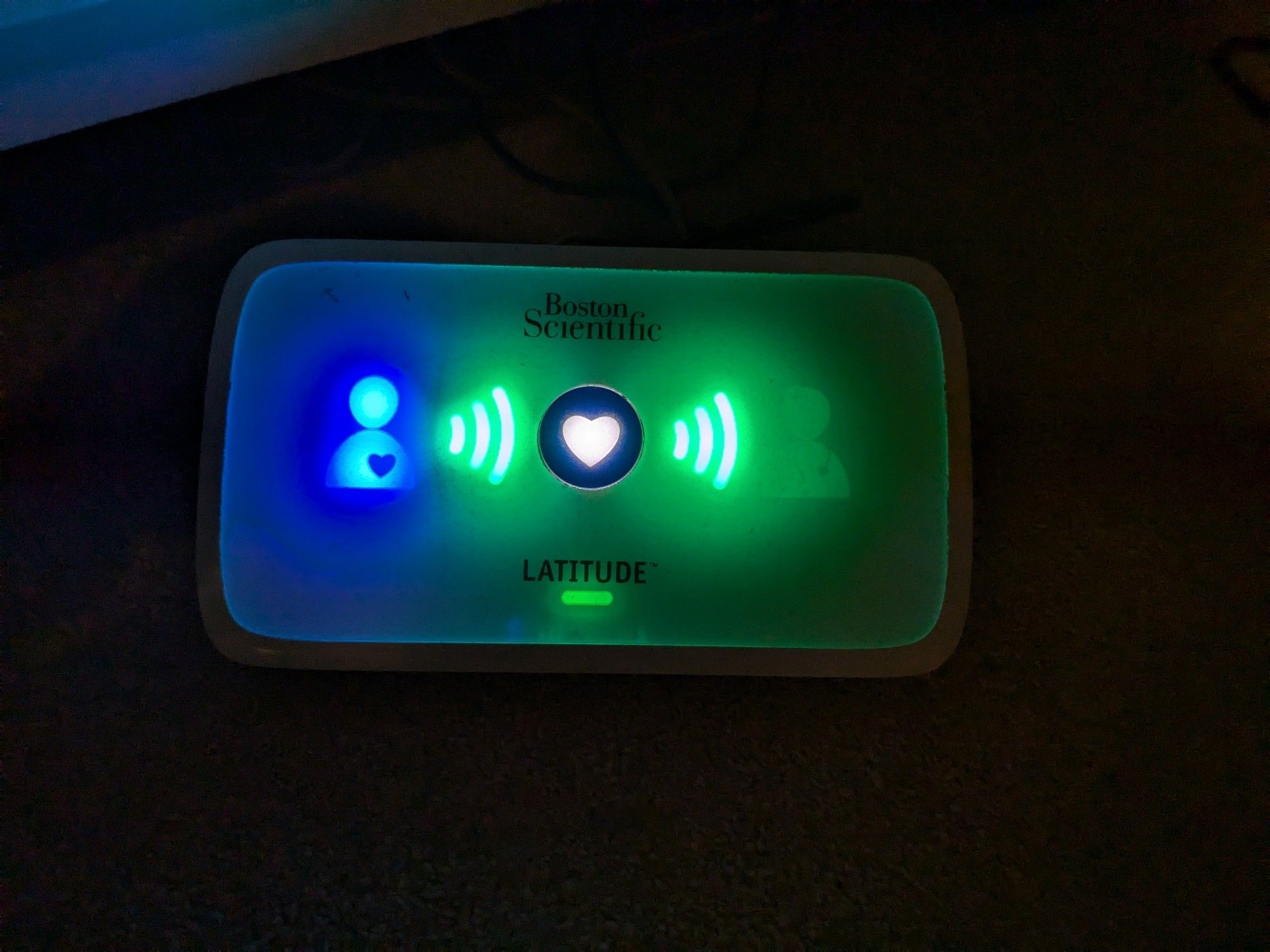 Picture of ICD Remote monitoring device. It is a white box with, from left to right illuminated graphic of a torso and head with a heart in it. Then a wireless logo, then a button, then another wireless logo and finally unilluminated a graphic of the tose and head of someone with a cross on them, to indicate a clinician.  Below are the words Latitude and a green light.