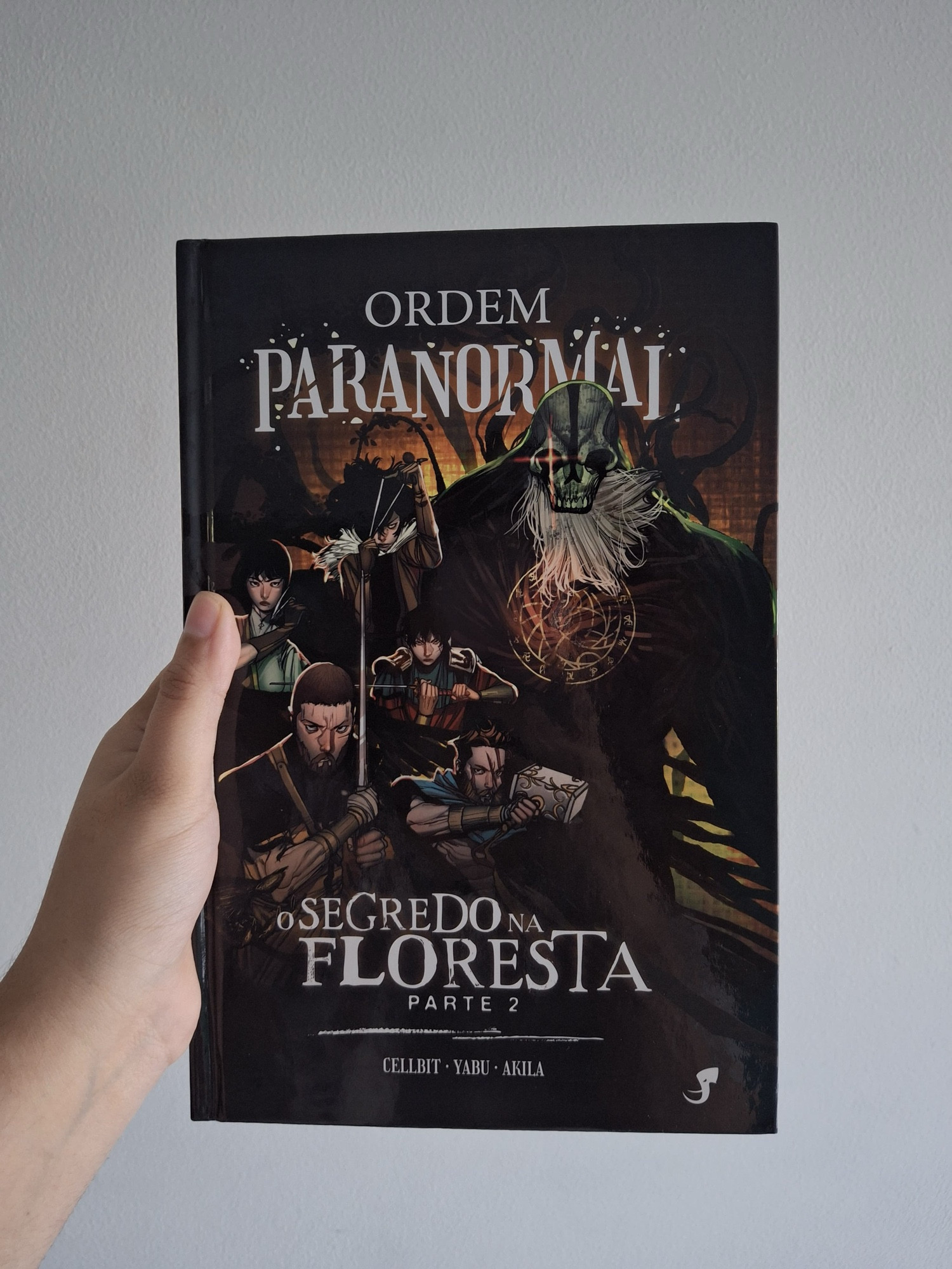 Capa da HQ de O Segredo Na Floresta - Parte 2. Do lado esquerdo vemos Elizabeth webber, mulher branca e morena segurando uma foice; ao lado, vemos César Cohen, homem branco e moreno segurando um estilingue; embaixo, vemos Thiago Fritz, homem branco e moreno segurando uma espada; ao lado, vemos Joui Jouki, homem japonês e moreno segurando uma espada; embaixo, vemos Arthur Cervero, homem branco e moreno segurando um martelo. Do lado direito, vemos o Deus da Morte. O fundo é branco e uma mão branca segura a HQ.