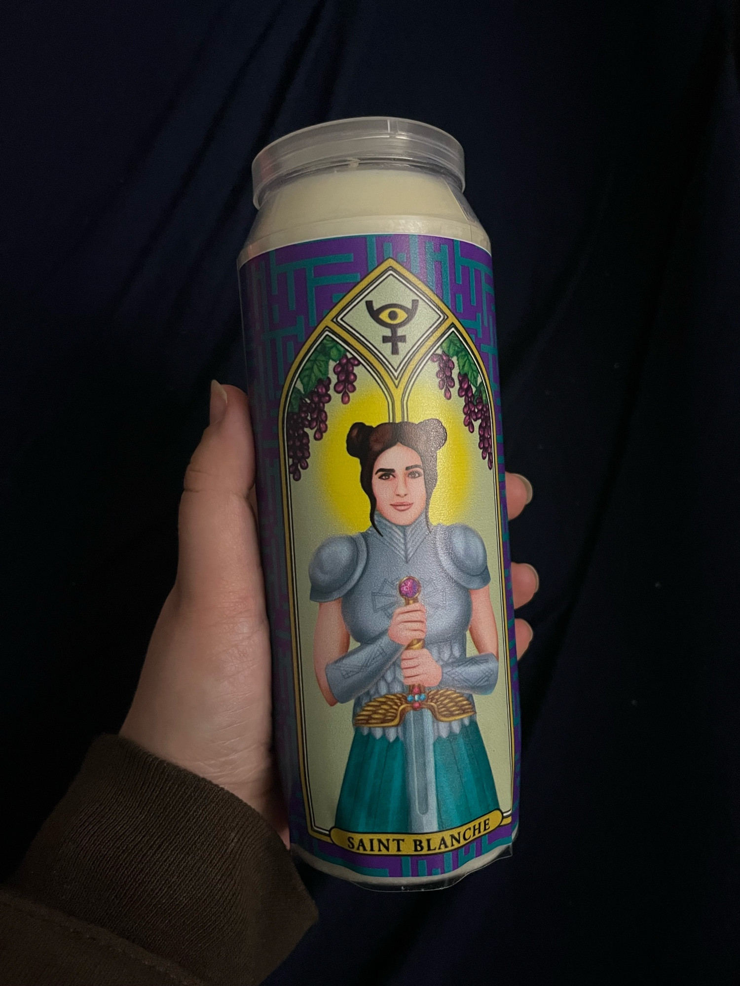 prayer candle depicting a pale skinned woman wearing armor and holding a sword. under her it says “SAINT BLANCHE.”