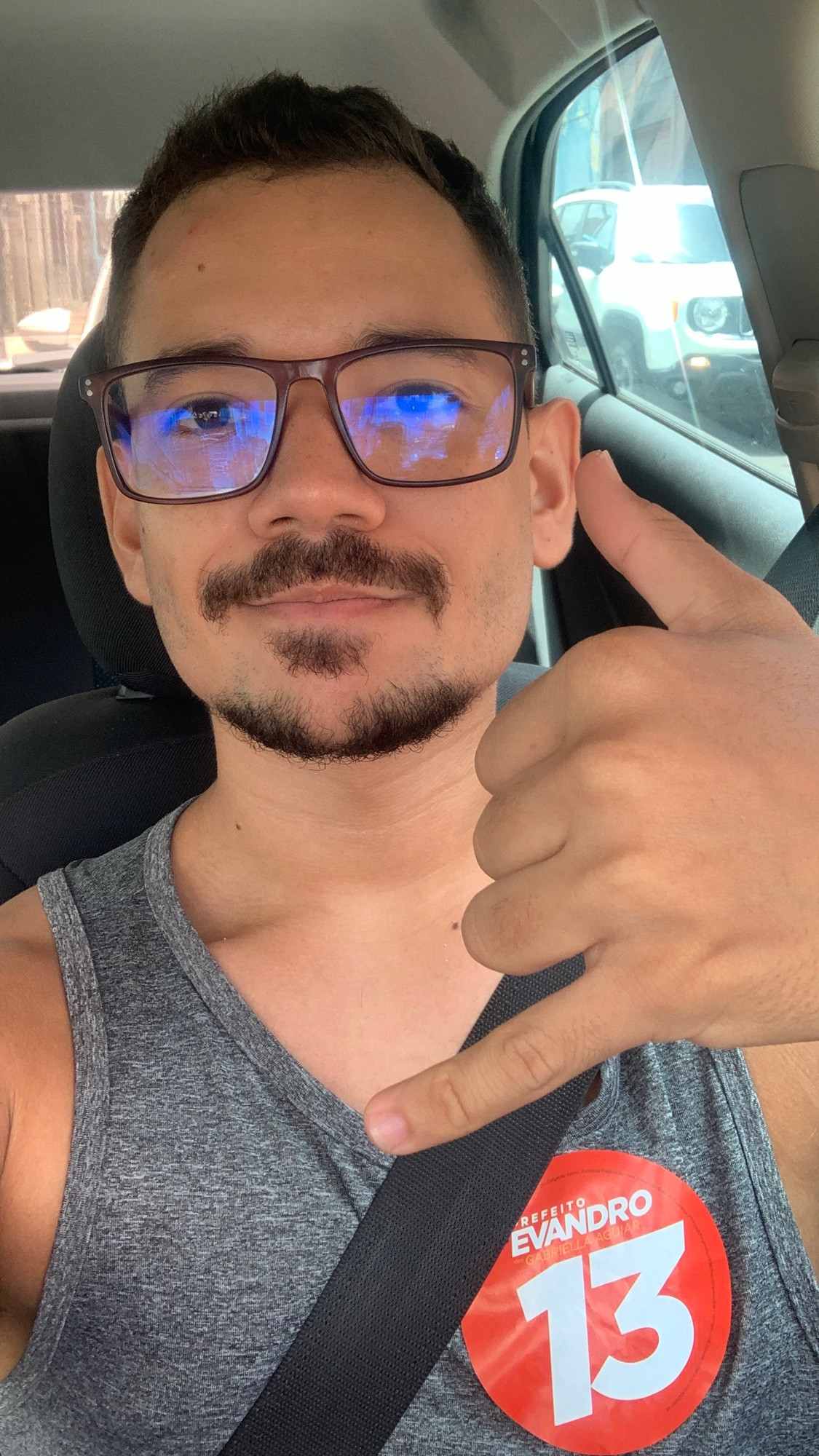 Selfie minha dentro do carro de regata, óculos de grau e um adesivo Evandro 13, fazendo hang loose