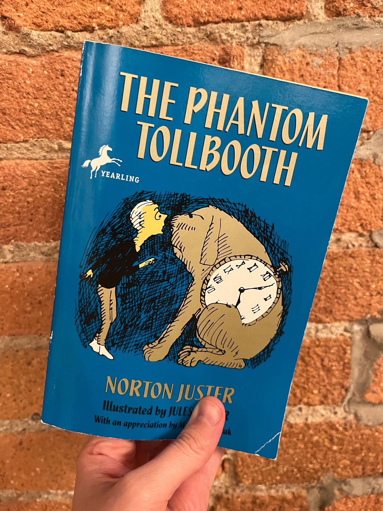 A copy of THE PHANTOM TOLLBOOTH by Norton Juster held against a brick wall.