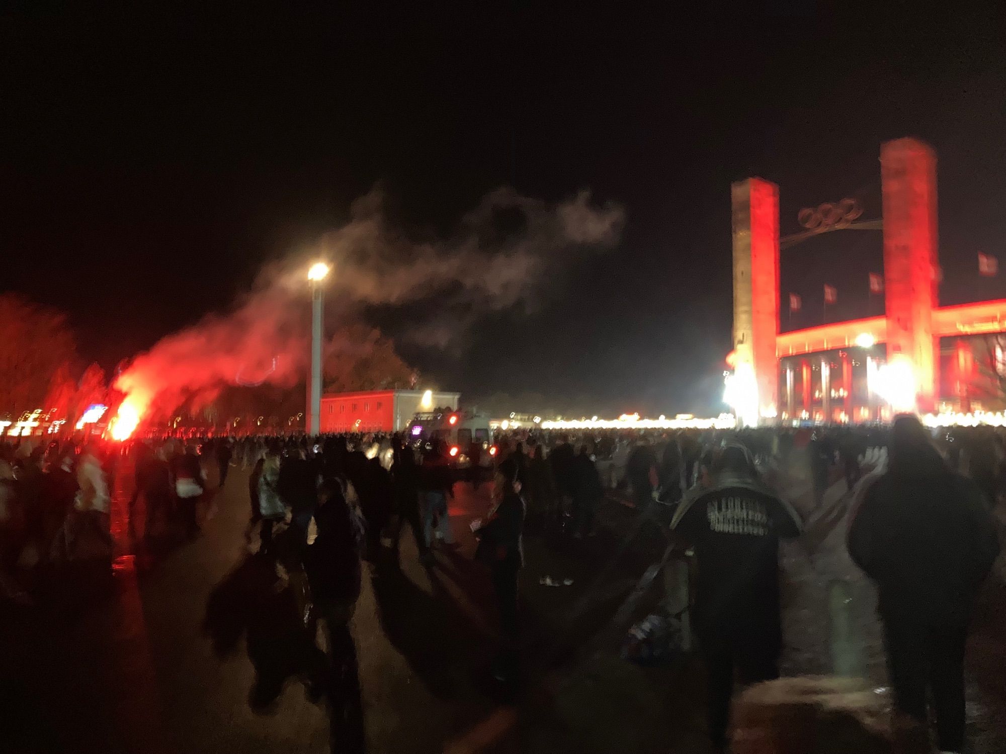 Auf der rechten Seite das Berliner Olympiastadion in rot mit roter Pyro von der linken Seite. Im Vordergrund dunkle Fans darüber Rauch im schwarzen Nachthimmel.